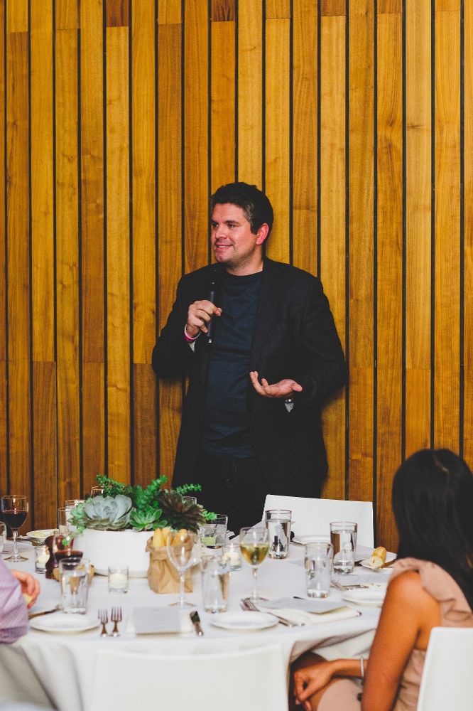 Andrew Casale holding a microphone and speaking to colleagues seated around a dinner table