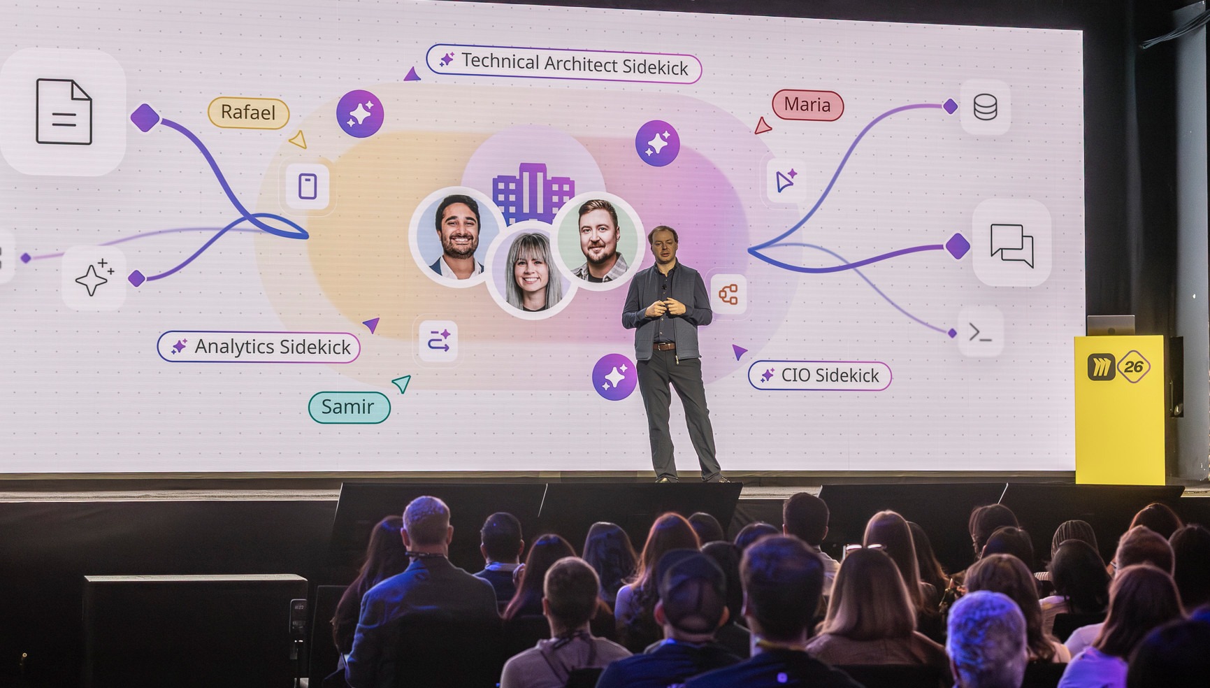Andrey Khusid speaking onstage at Miro Canvas 26 in front of a screen showing AI Sidekicks and team members