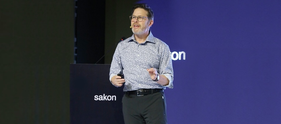 Andy Goorno speaking onstage beside a Sakon lectern at the company’s Pune gathering