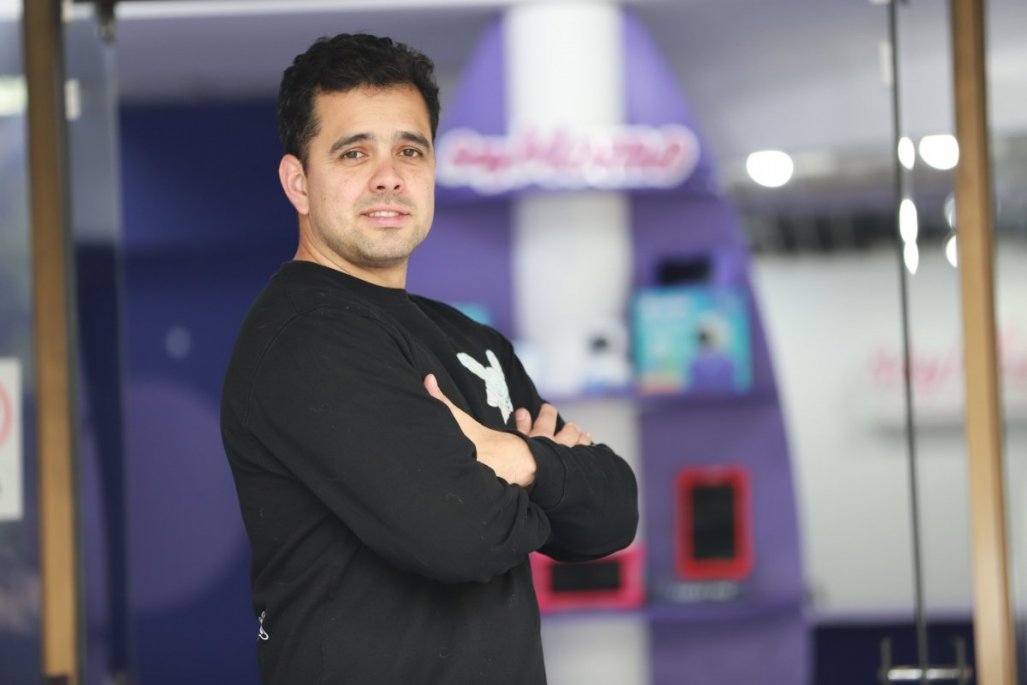 Aníbal Madrid standing with folded arms at SoyMomo, with purple branding and devices behind him