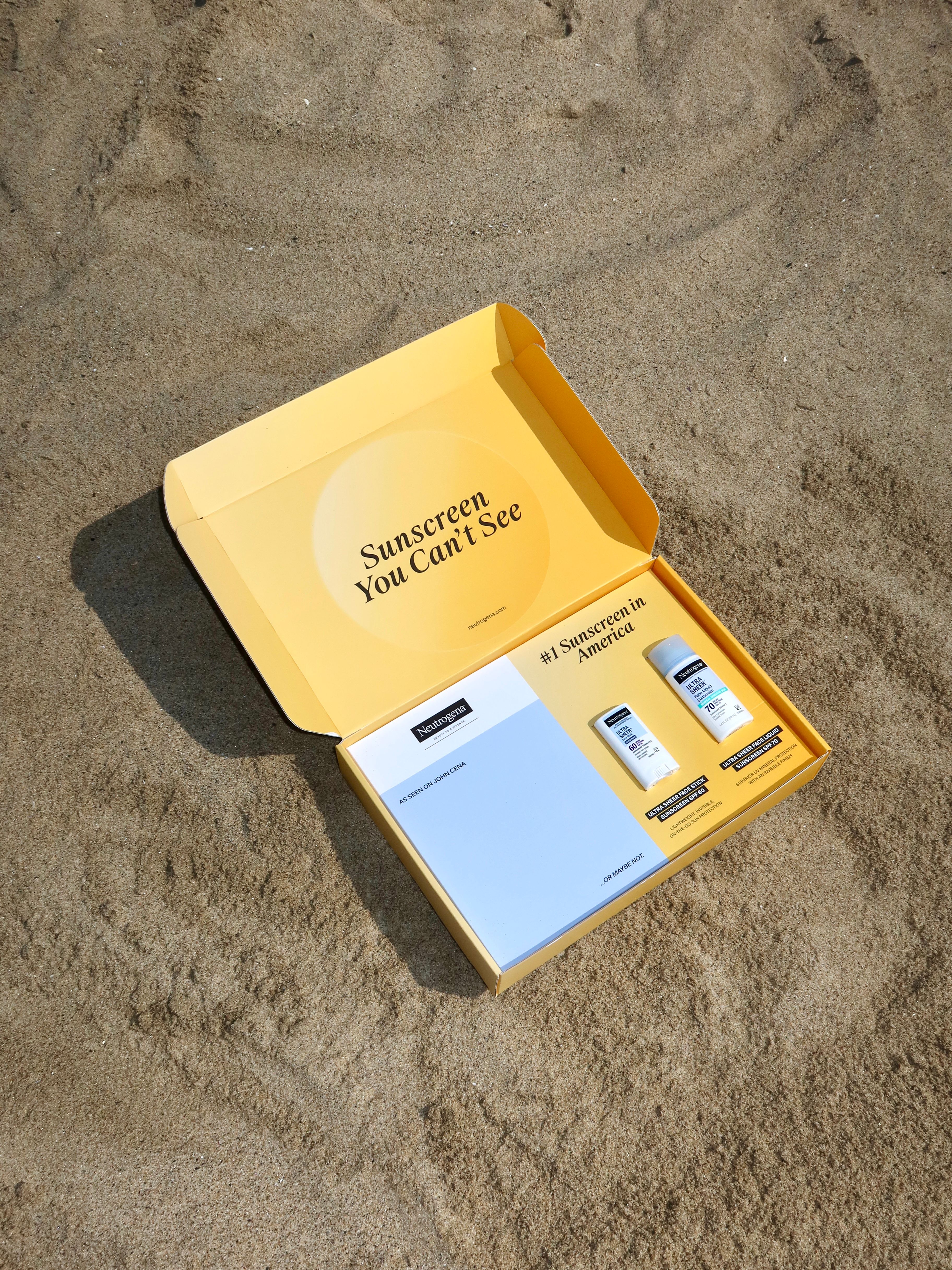 Open yellow Neutrogena Ultra Sheer gifting box on sand, holding sunscreen and a campaign insert