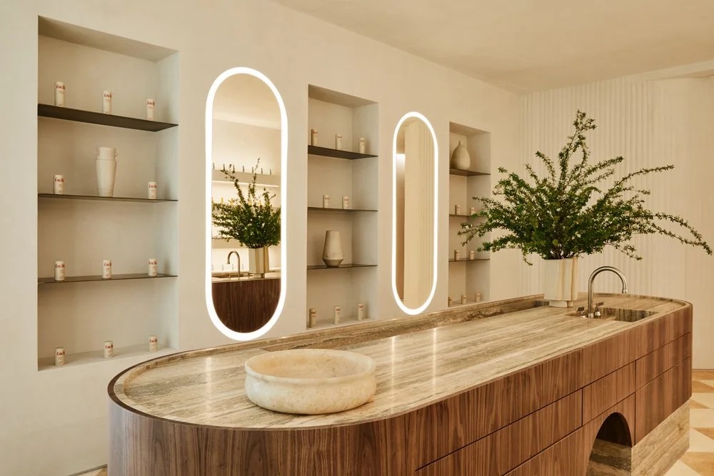 Facile interior with illuminated oval mirrors, warm wood cabinetry and skincare products on shelves