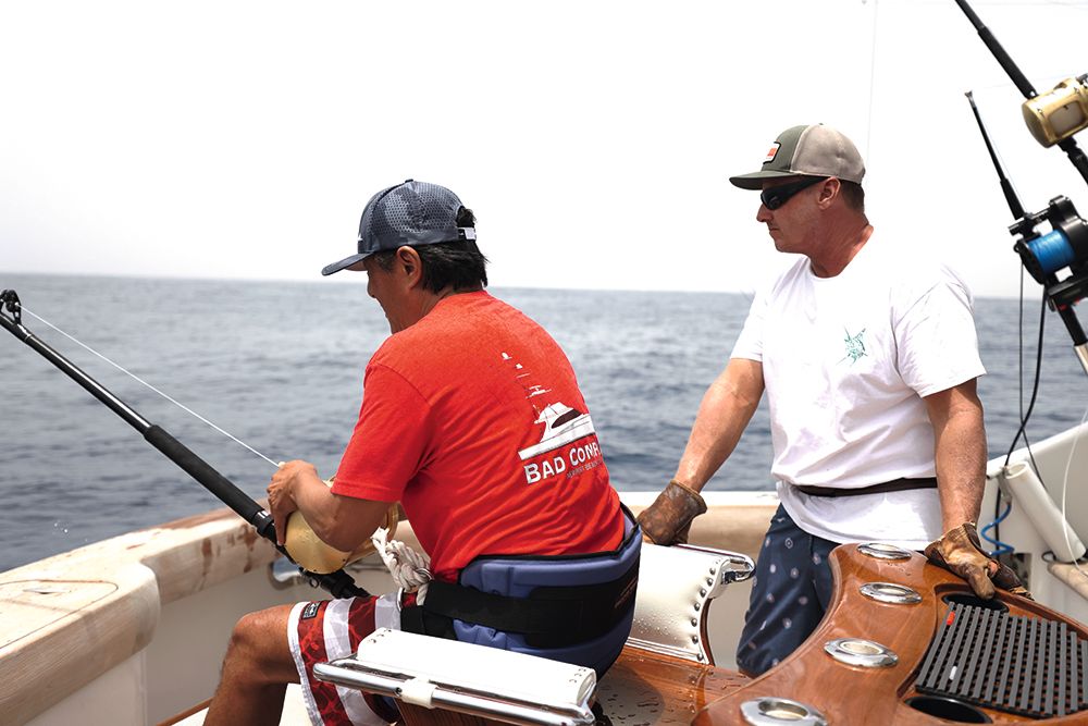 Anthony Hsieh in a red Bad Company shirt working a fishing rod aboard a boat, with a crew member beside him.