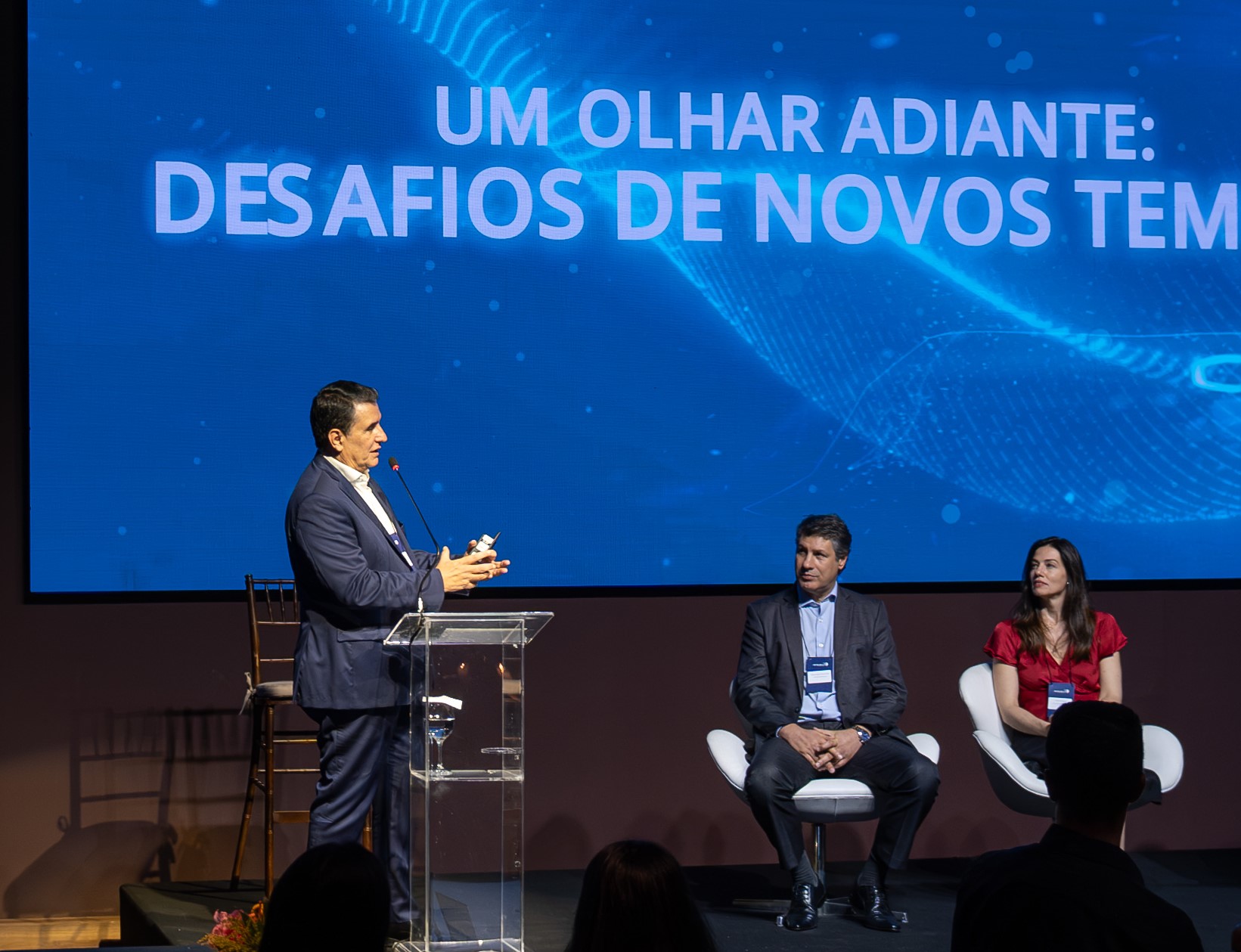 Antonio Nasser seated onstage at ABIMED’s annual meeting, with a speaker at the lectern and another participant beside him.