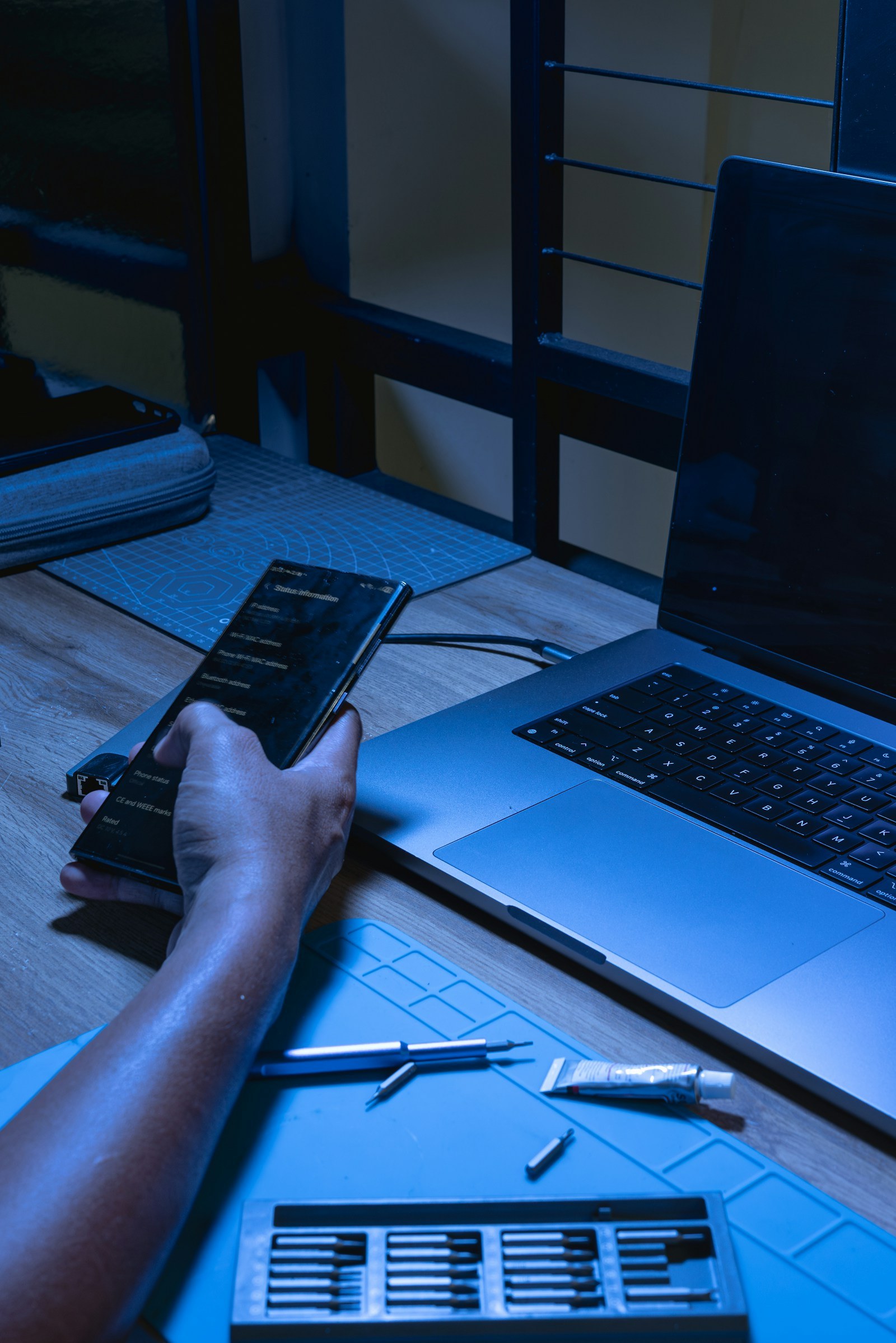 A person using a phone beside a laptop in a blue-lit workspace