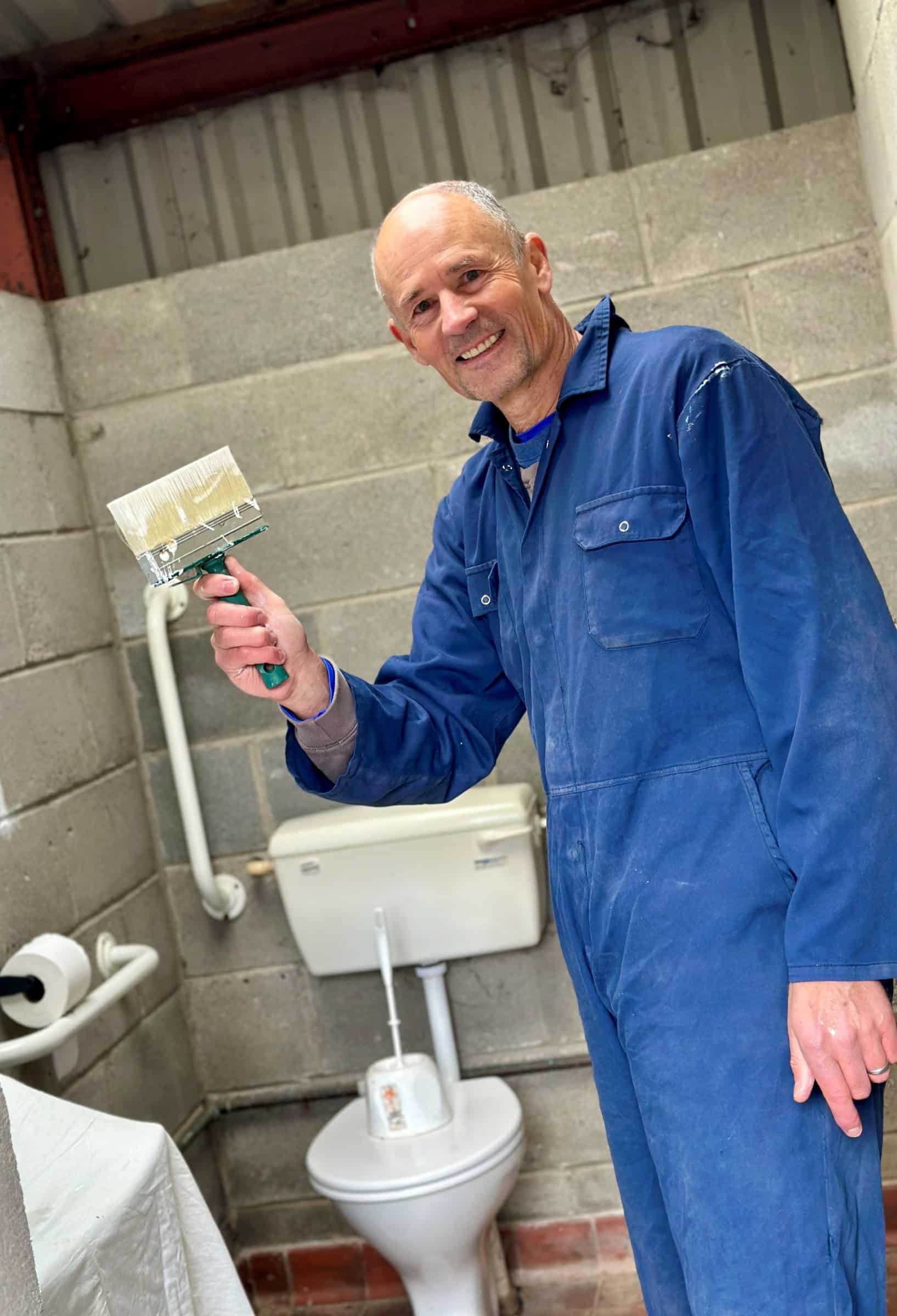 A smiling volunteer in blue overalls holds a paintbrush during decorating work, in a photograph published by Applicable.