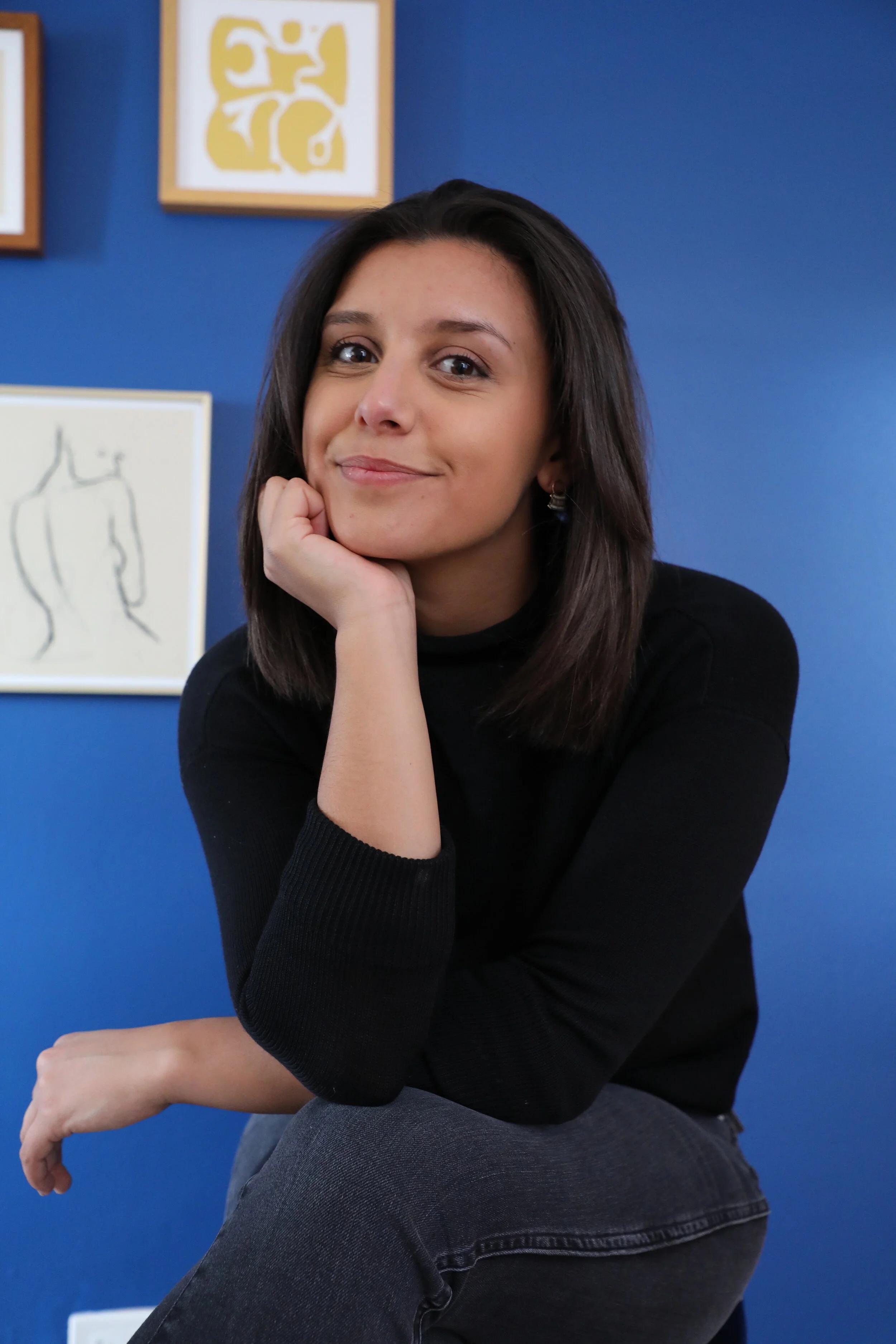 Ariela Safira seated in front of a blue wall with framed artwork
