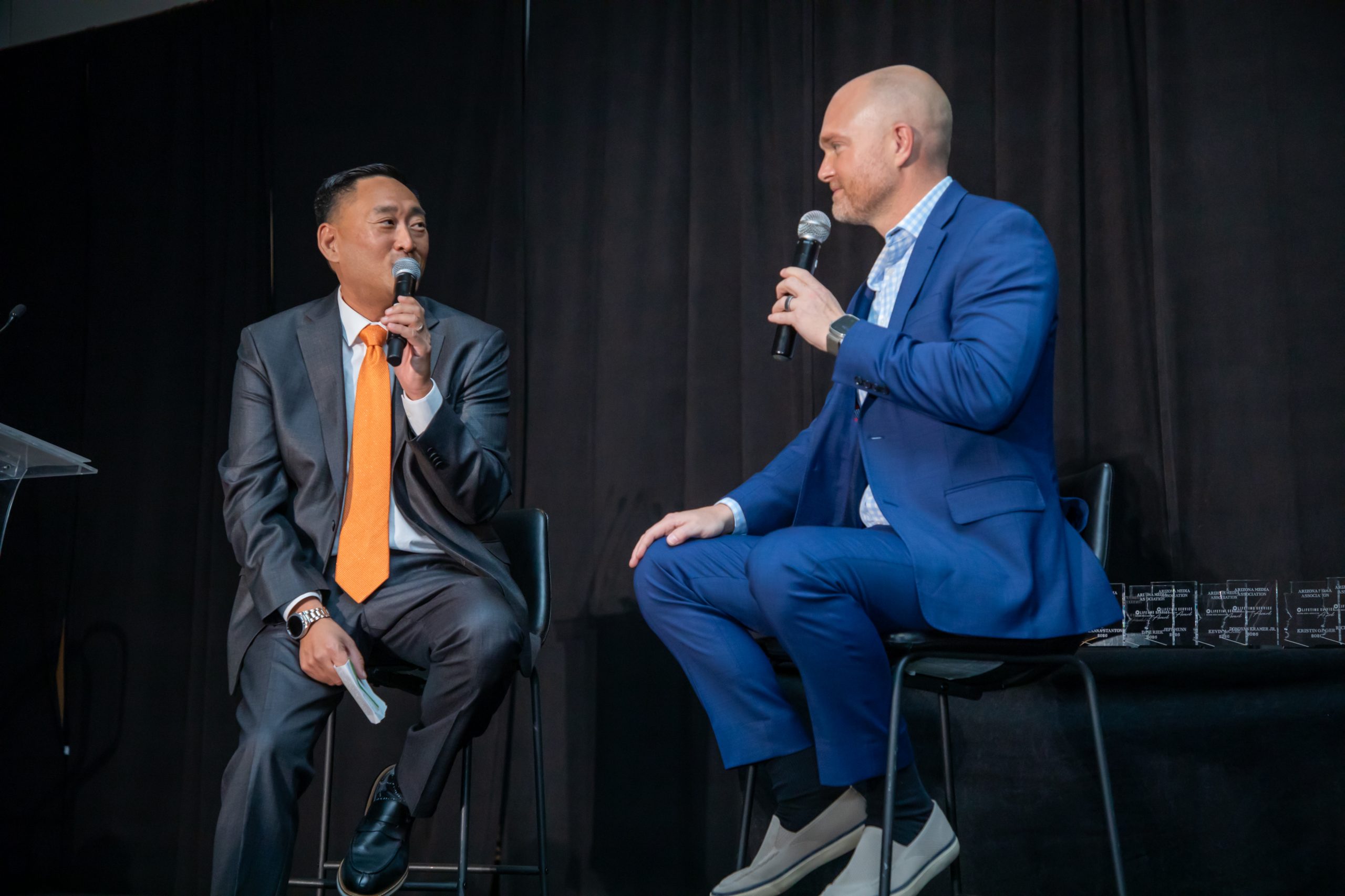 Two Arizona media professionals speaking onstage with microphones at an association event