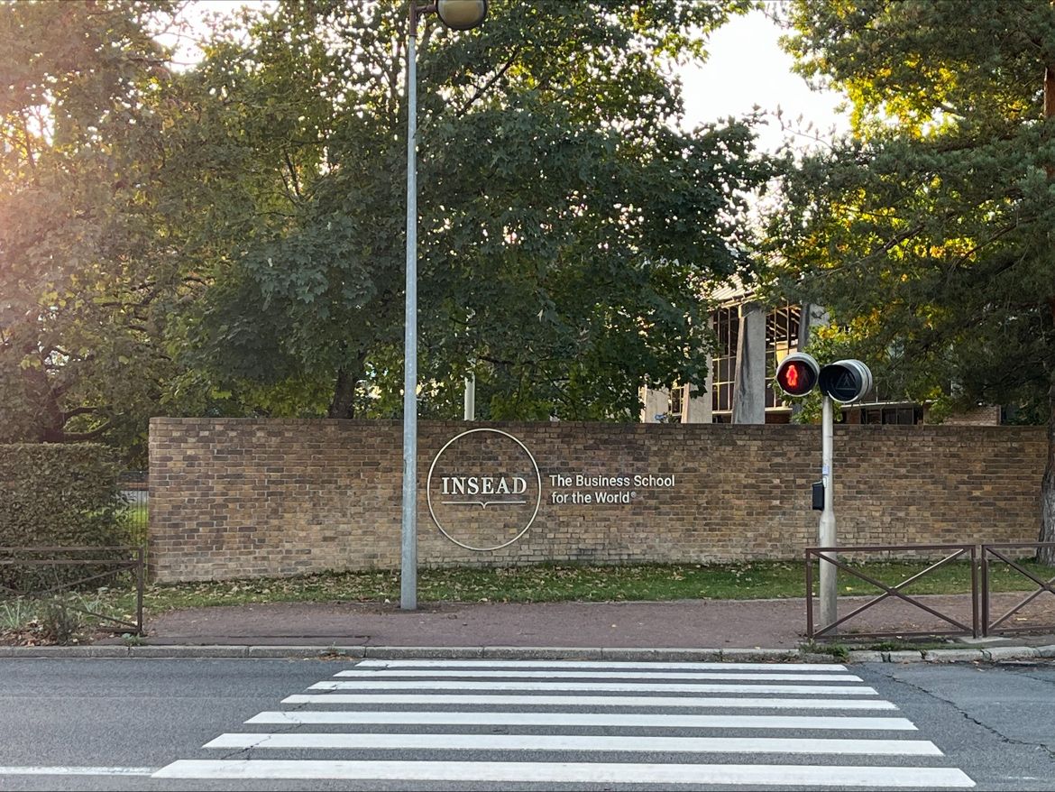 Tree-lined entrance to INSEAD, with its name on a brick wall and a pedestrian crossing in front.