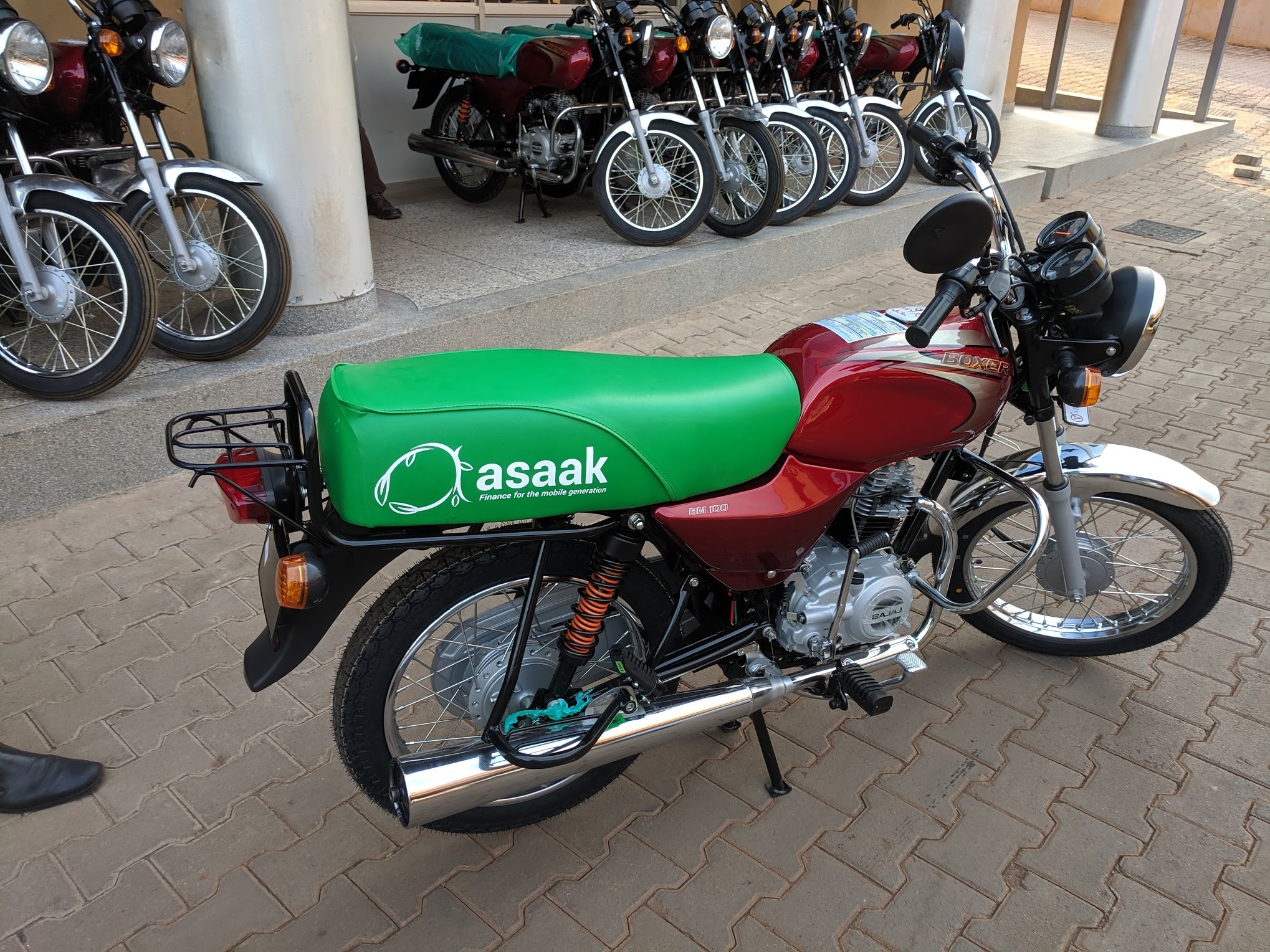 A red motorcycle with a green Asaak-branded seat cover, with more motorcycles parked behind it