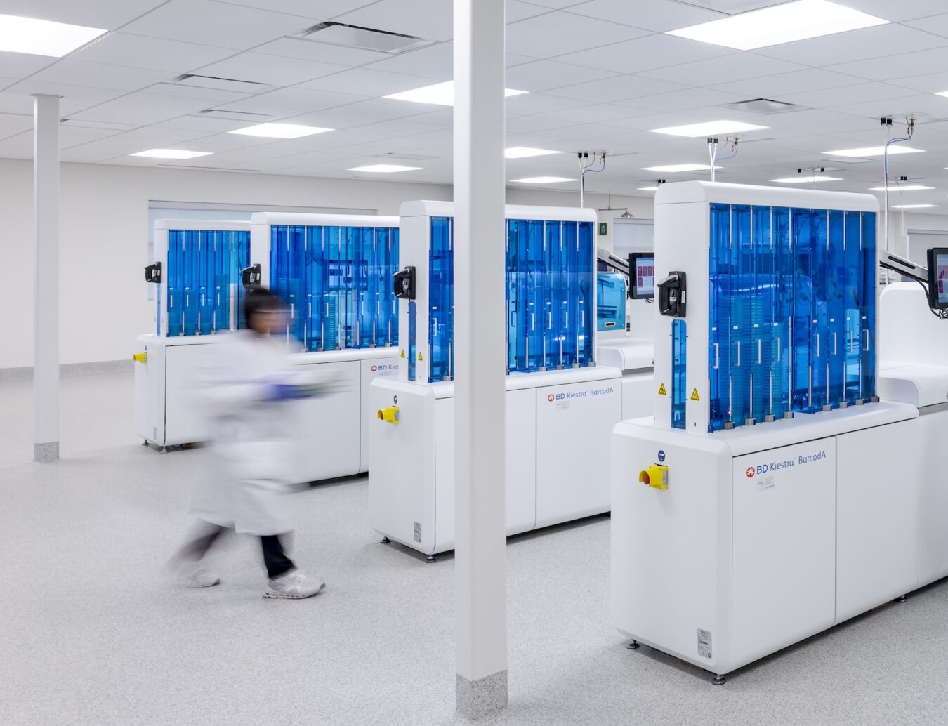 Automated laboratory equipment with blue enclosures and a staff member walking between instruments