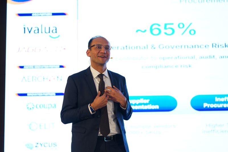 Ashutosh Dhar speaking on stage at a procurement summit, wearing a suit and a headset microphone