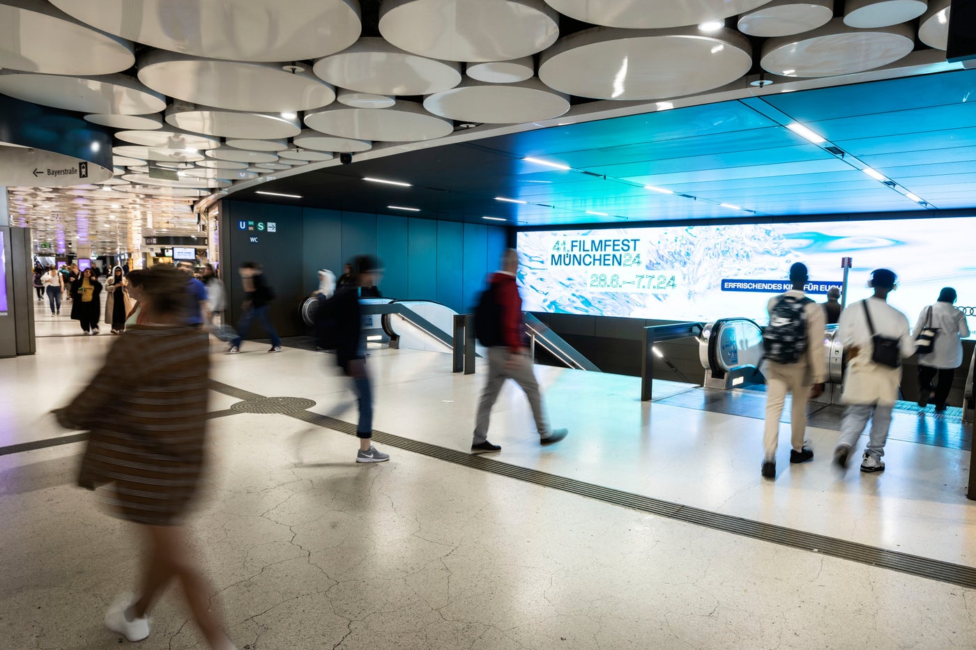 Commuters pass a bright FILMFEST MÜNCHEN 2024 display beside an escalator