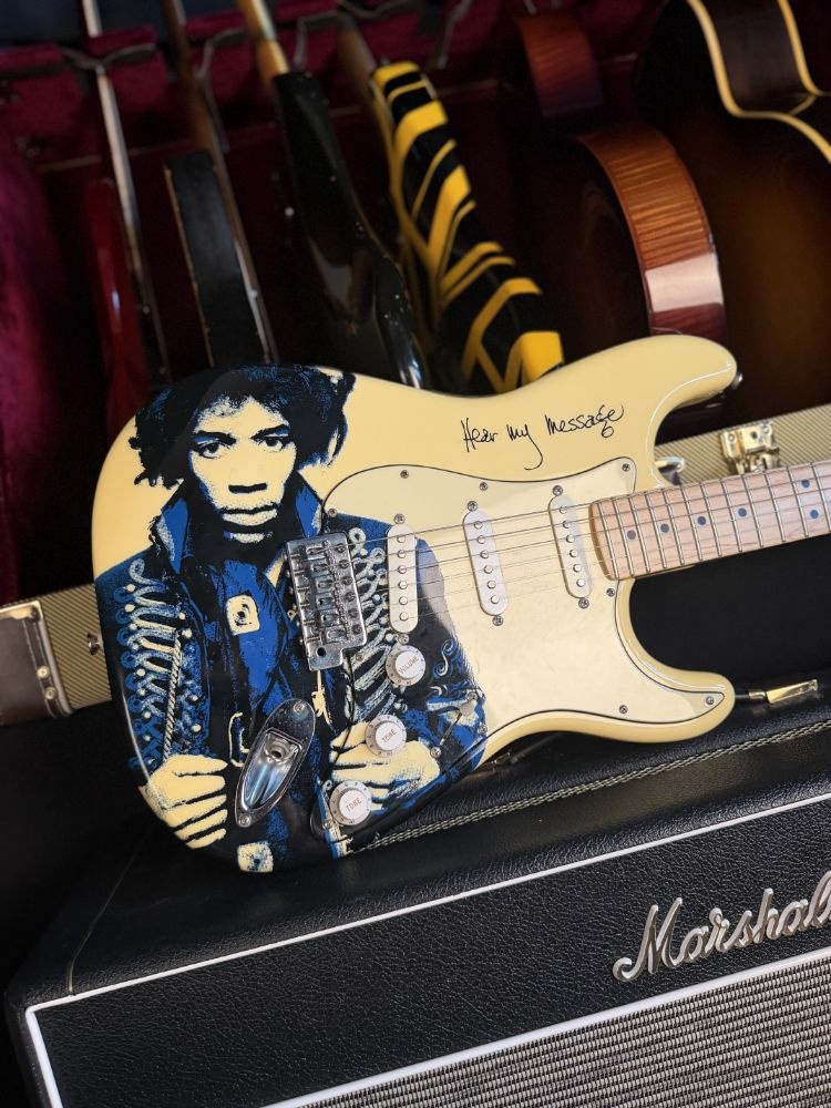 A cream electric guitar decorated with a portrait, resting on an amplifier among other guitars