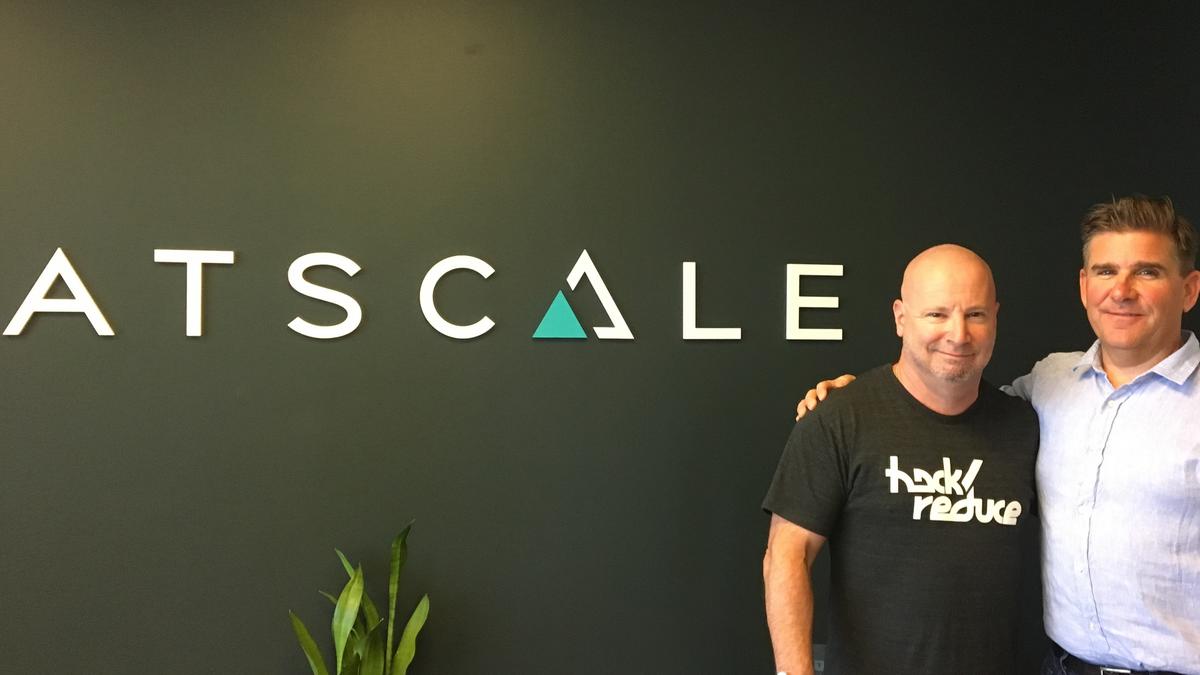 AtScale leaders Chris Lynch and Dave Mariani standing beside an AtScale wall logo