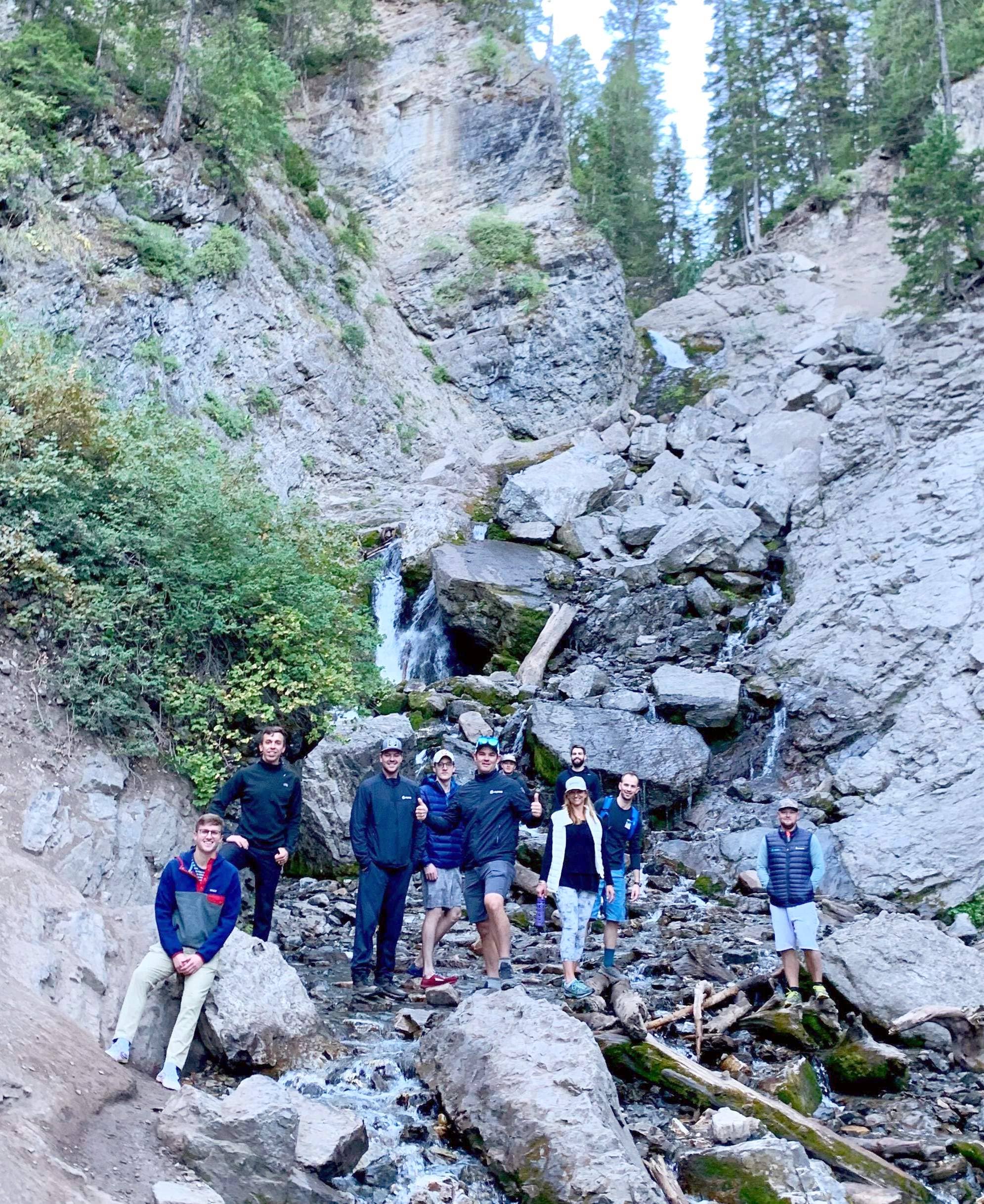 Aumni team members standing on a rocky trail beside a waterfall in a historical team photograph