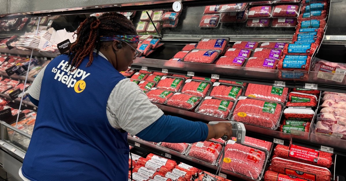 A Walmart associate uses a handheld reader beside packages in a refrigerated meat display.
