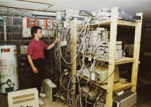 Archival photograph of Netaxs equipment in Wyndmoor, with cable-filled wooden shelving, computers and a person standing beside it