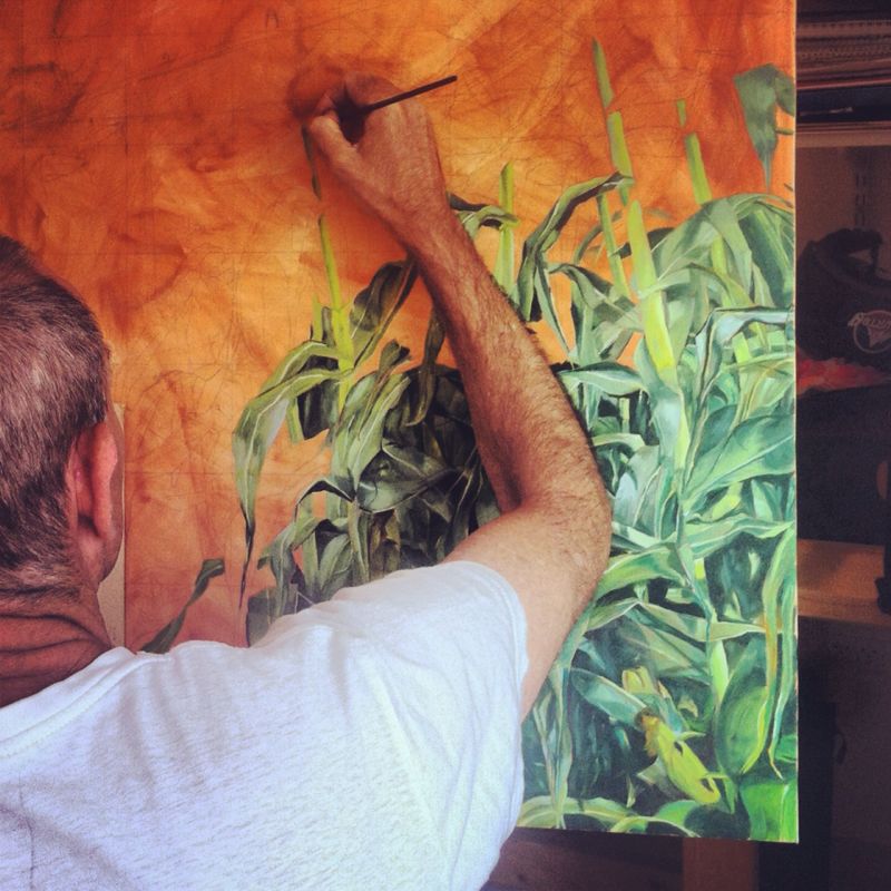 A brush held above a canvas with green plants against an orange ground, from Jacoby’s painting practice