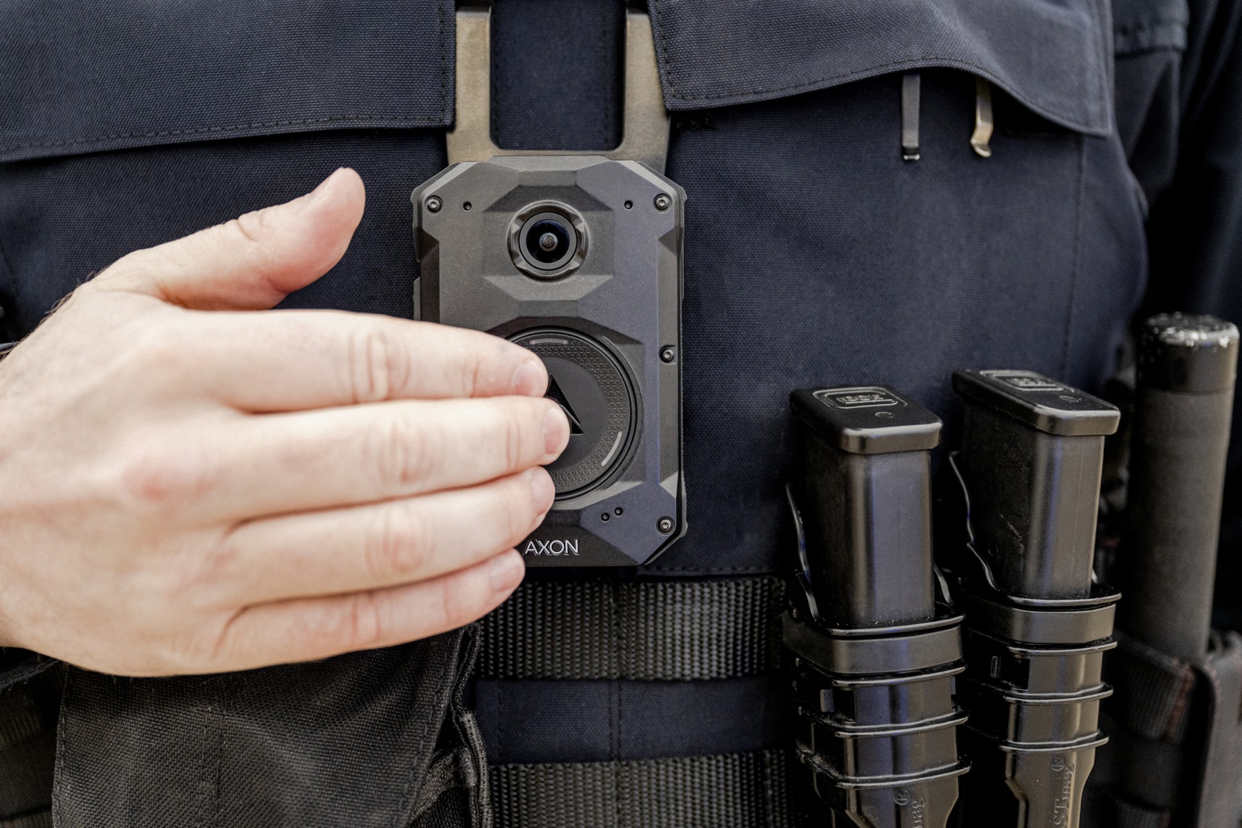 A hand operates an Axon Body 4 camera mounted on a police uniform