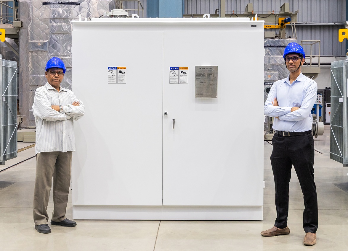 Two people wearing blue hard hats stand beside a large Ayr Energy electrical cabinet on a factory floor