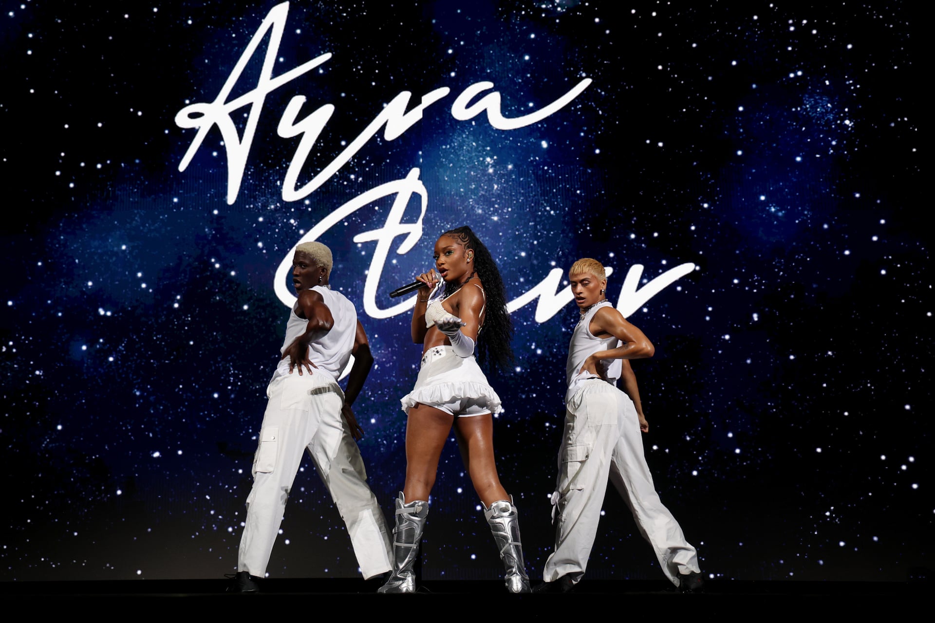 Ayra Starr performing onstage in white with two dancers against a starry backdrop