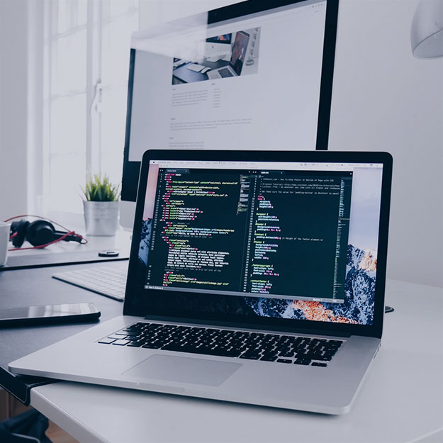 An illustrative workstation photograph from the company’s website, showing code on a laptop and a larger screen behind it