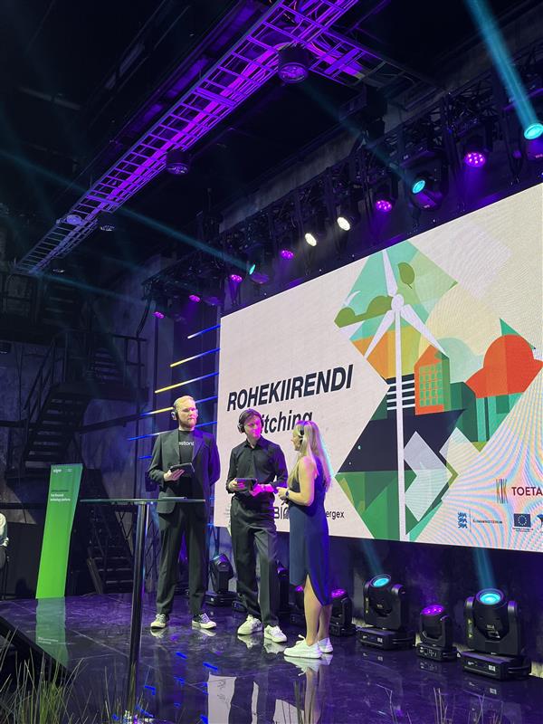 Three participants on a stage beneath a Rohekiirendi pitching display