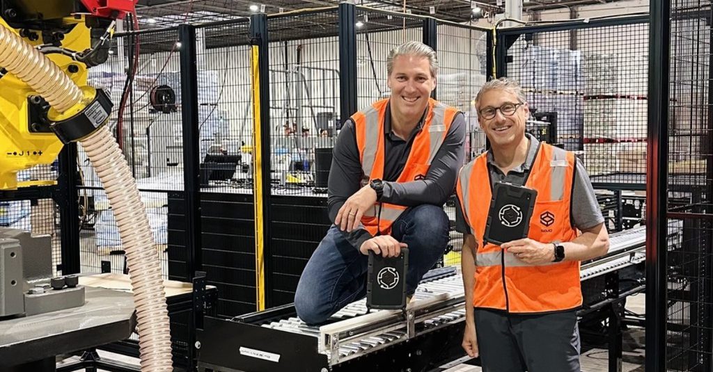 Barak Bar-Cohen and Steve Rubin in orange safety vests beside a Sojo robotic packaging line