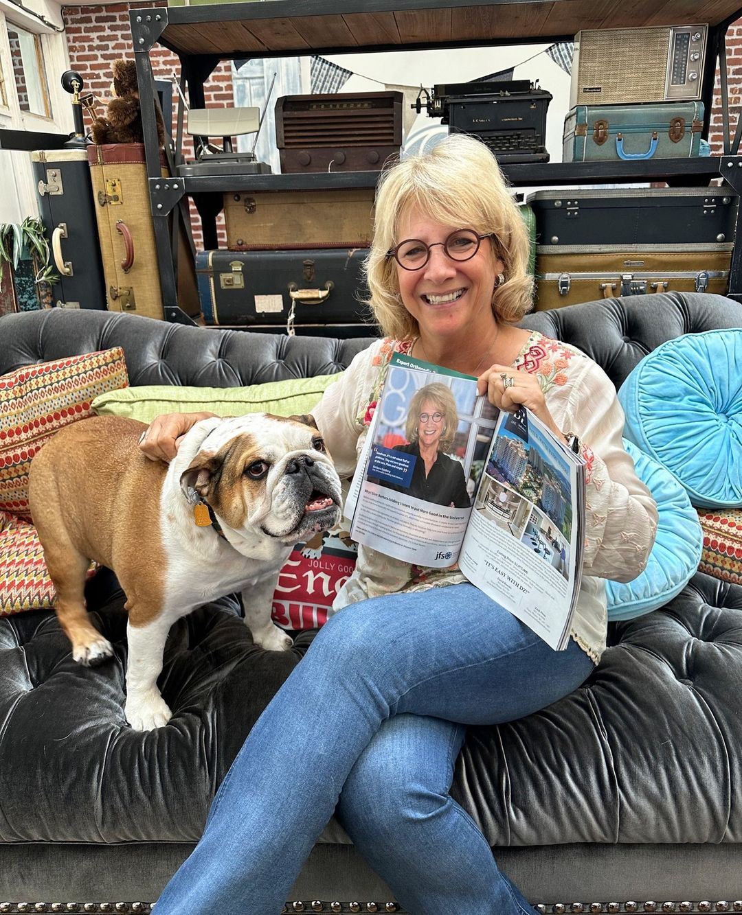 Barbara Goldberg holding open a magazine featuring her, with her dog Rosie beside her on a sofa