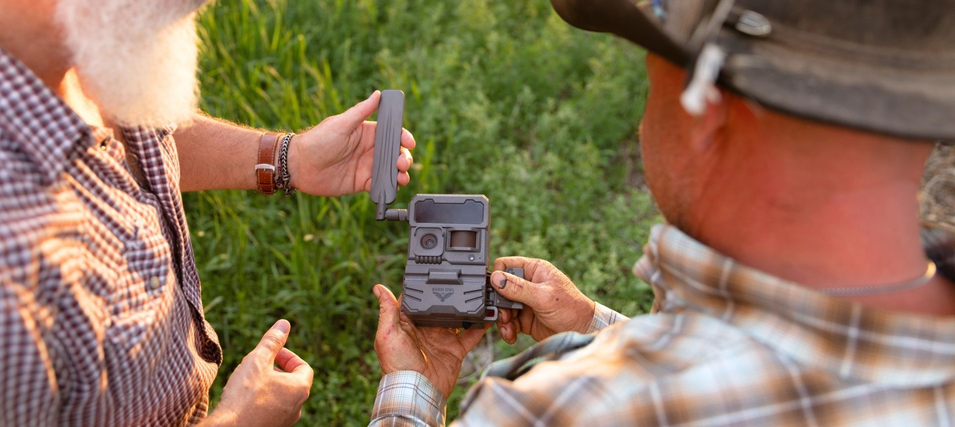 Two people in plaid shirts inspecting a Barn Owl camera outdoors in a green field