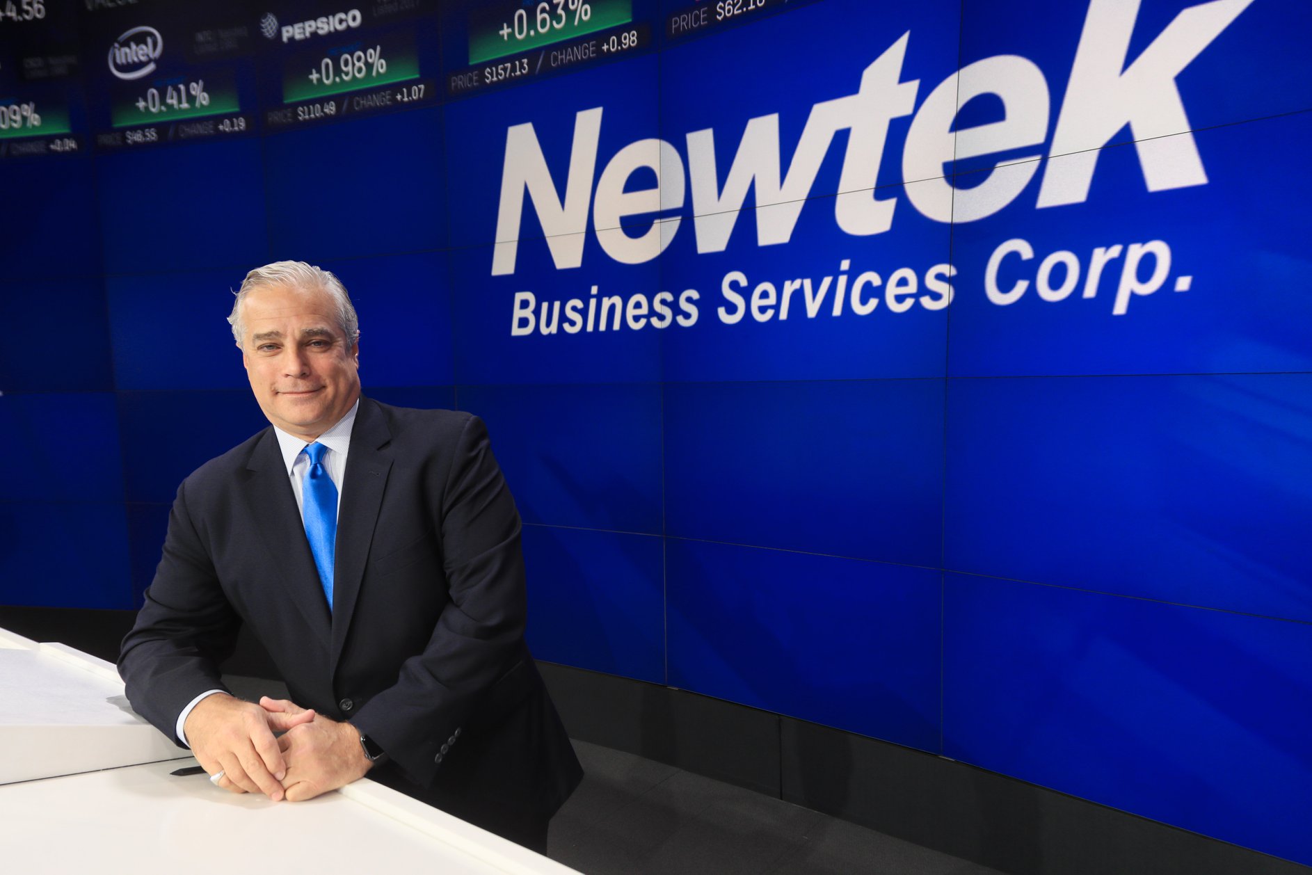 Barry Sloane seated beside a Nasdaq podium in front of a Newtek Business Services display
