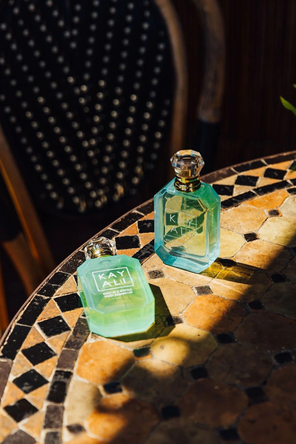 Two green KAYALI fragrance bottles on a patterned table in sunlight