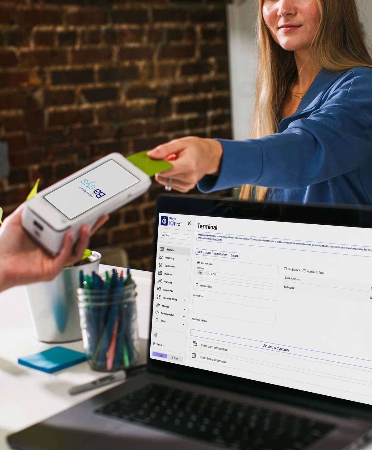A customer taps a payment card while the Basys iQ Pro+ terminal screen is open on a laptop