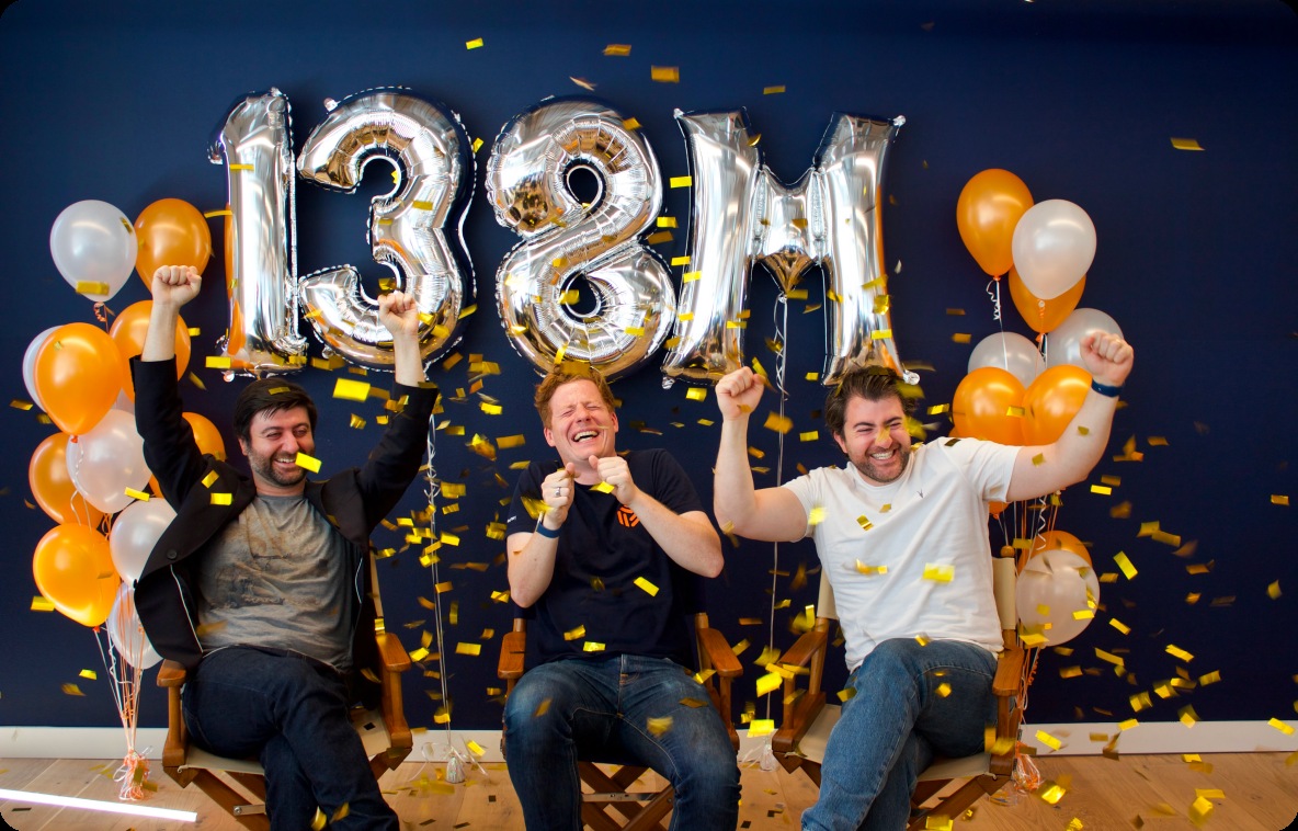 Three people celebrating beneath silver balloons spelling 138M at Beamery