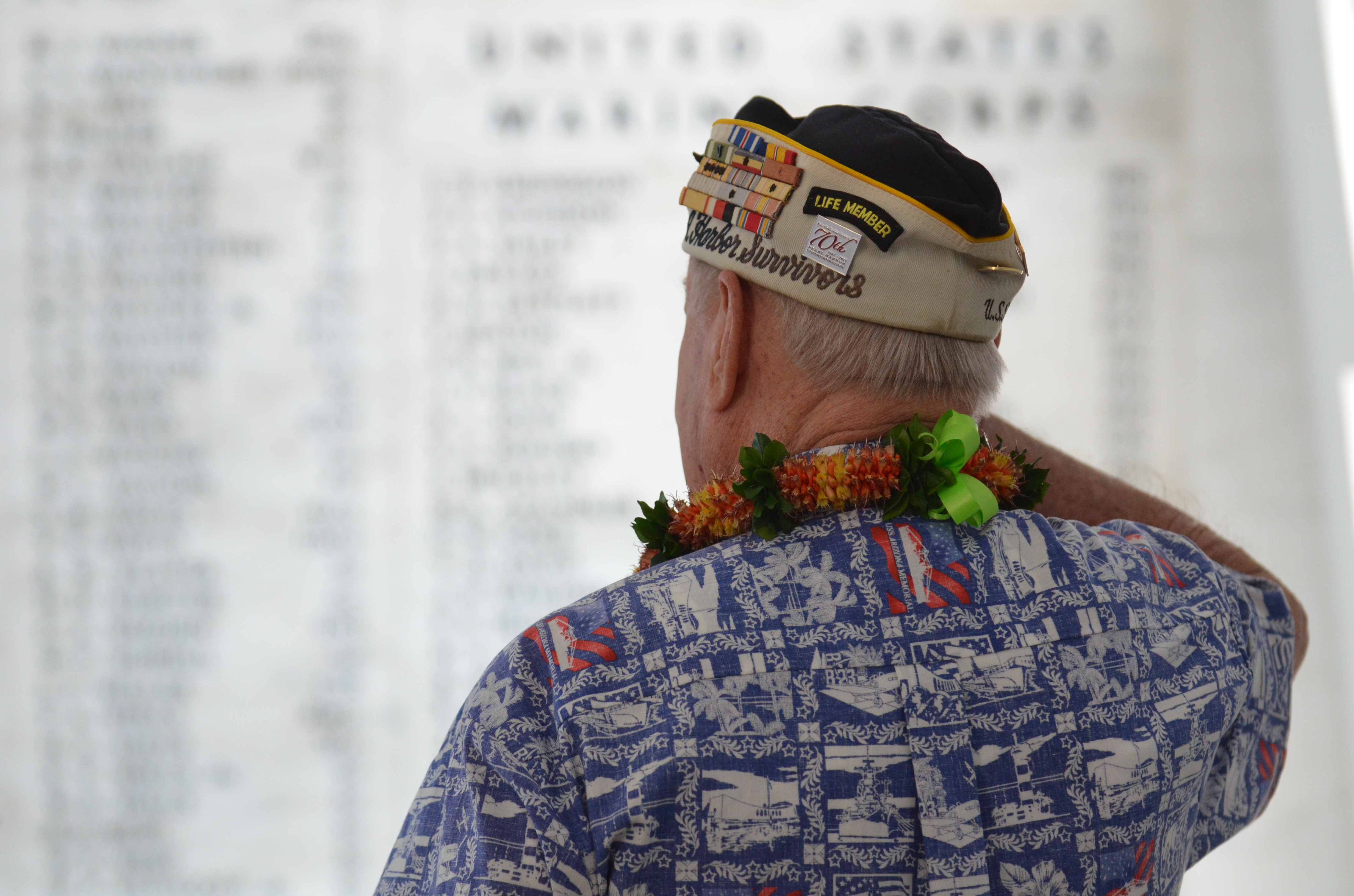 A Pearl Harbor survivor wearing a decorated veterans cap and lei looks toward a memorial wall