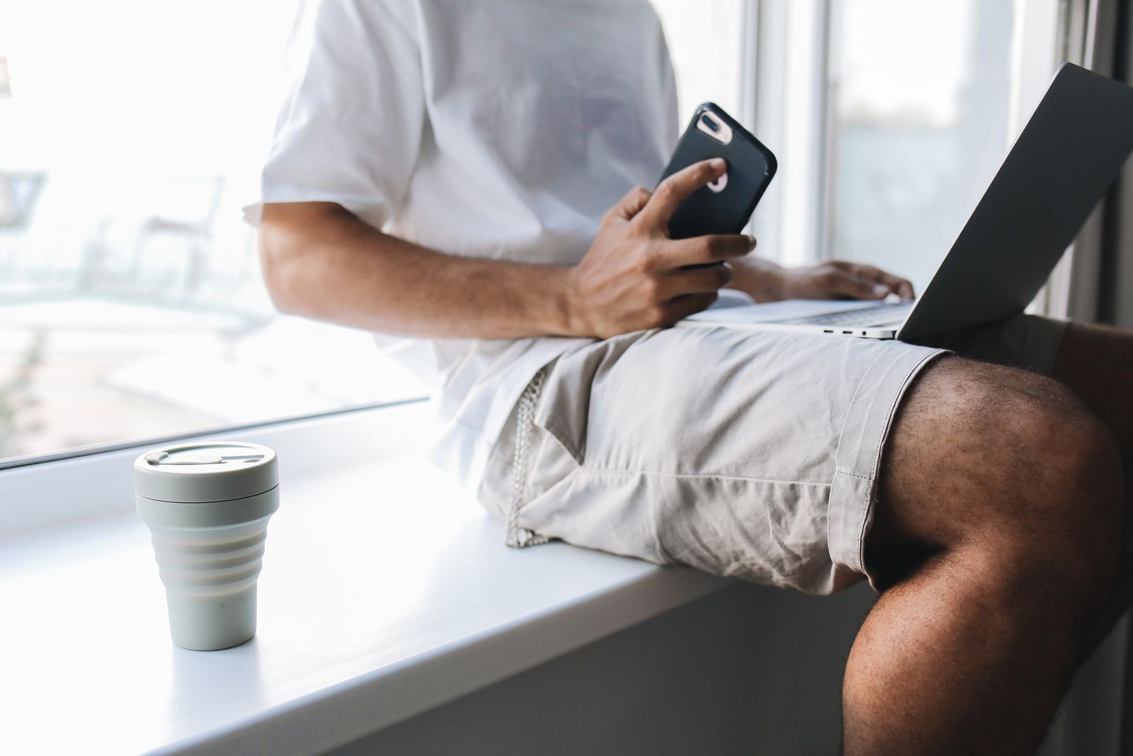 A person seated by a window using a laptop and phone