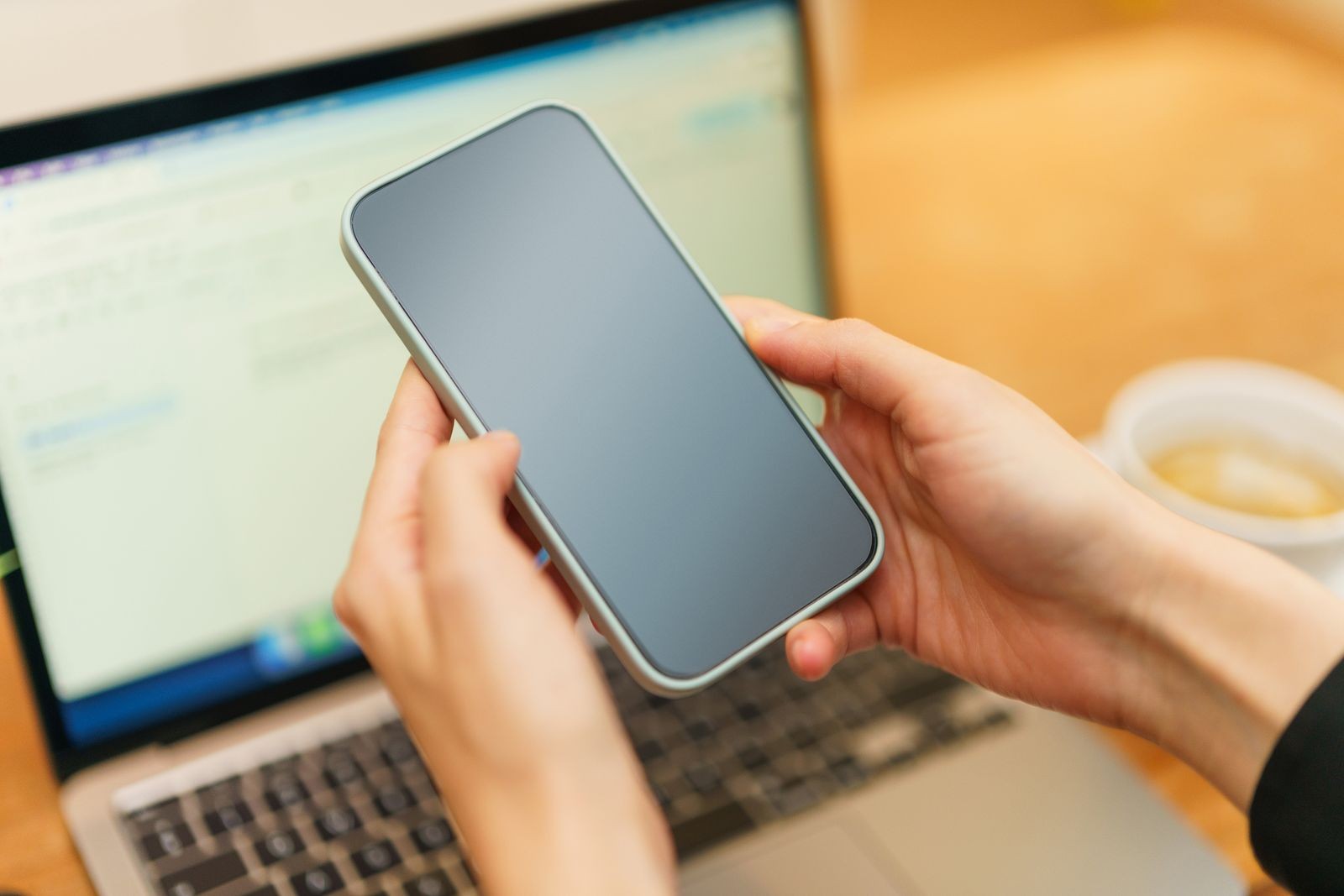 Hands holding a smartphone above a laptop beside a cup of coffee