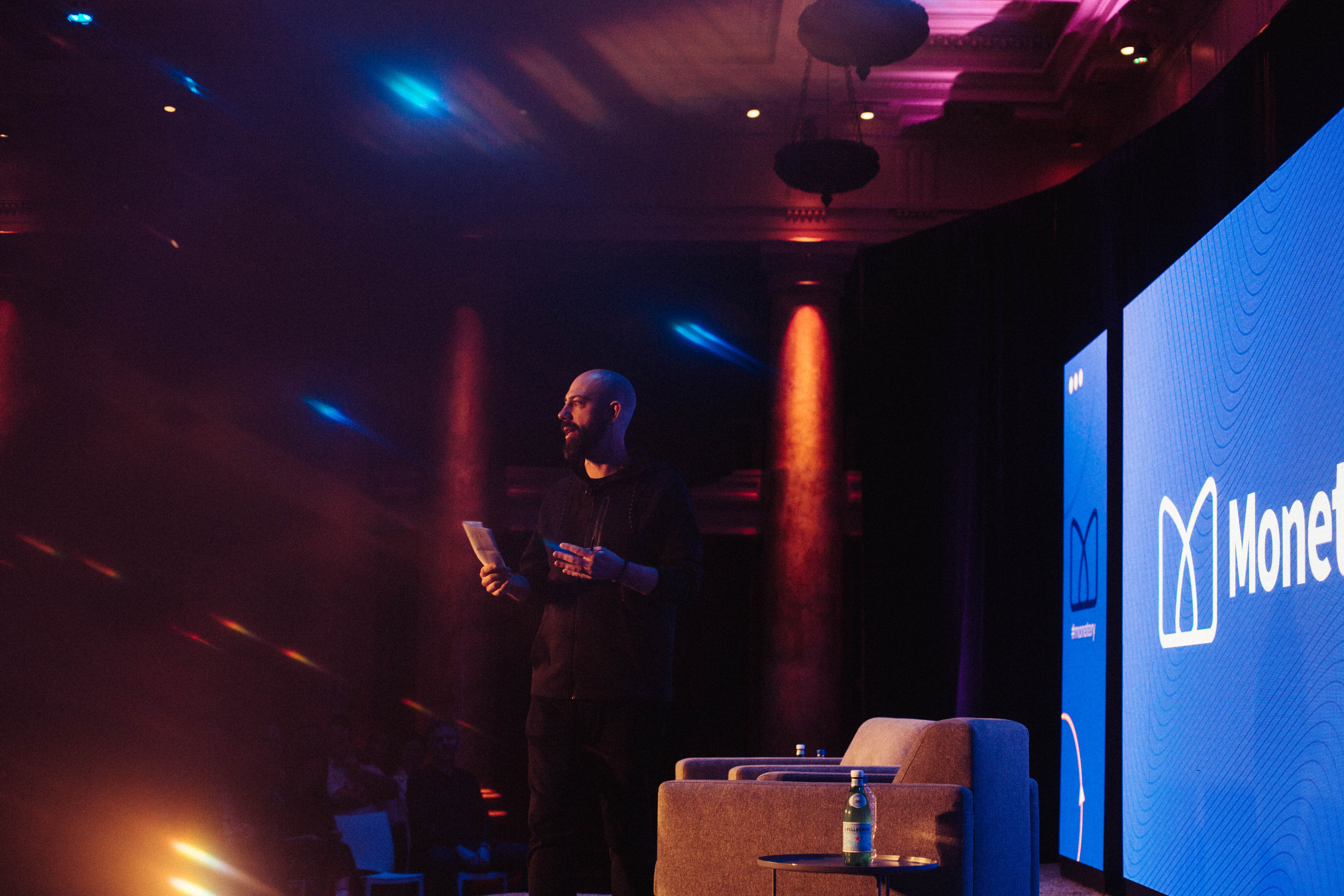 Ben Milne addressing attendees on the illuminated Monetery stage in Des Moines
