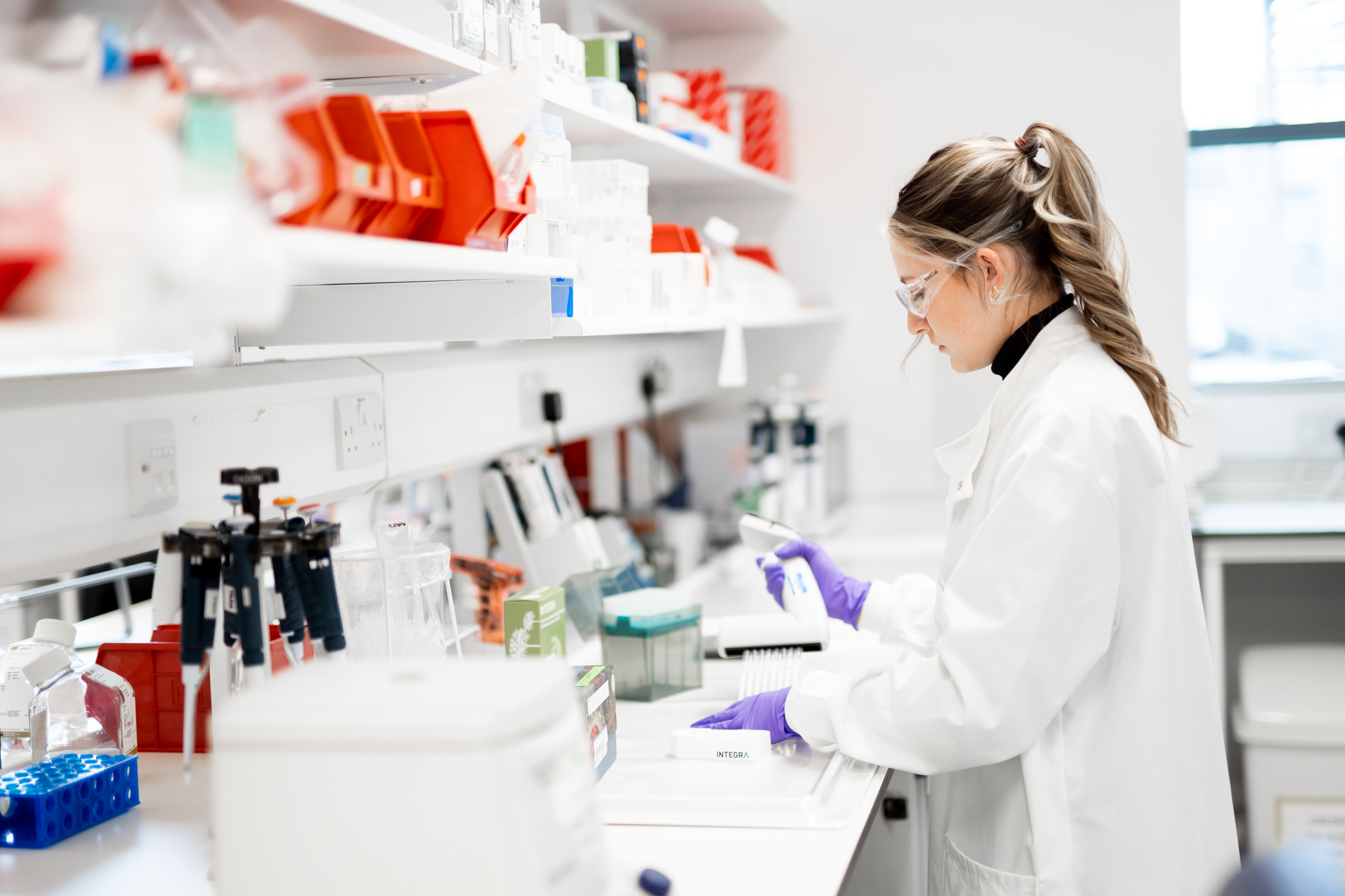 A researcher wearing safety glasses and purple gloves works at a bench in BenevolentAI’s Cambridge laboratory.