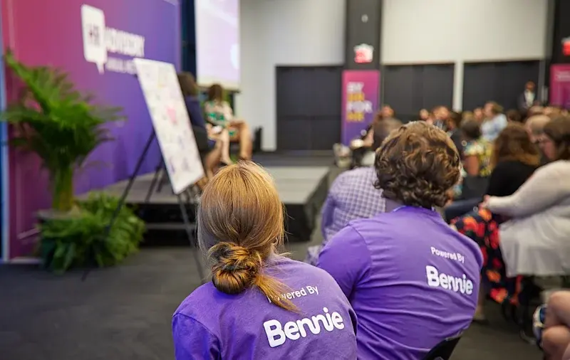 People attending an event, with two foreground attendees wearing purple Powered by Bennie shirts