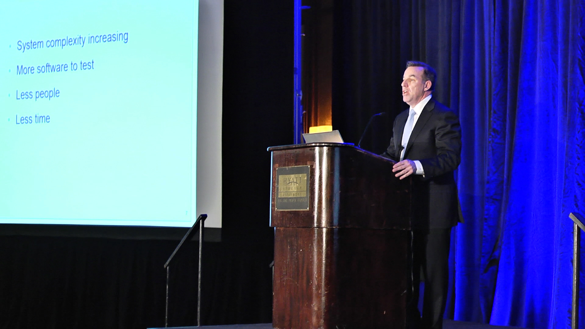 Bernie Gauf speaking at QUEST in 2014 beside a slide listing increasing complexity, more software, fewer people and less time