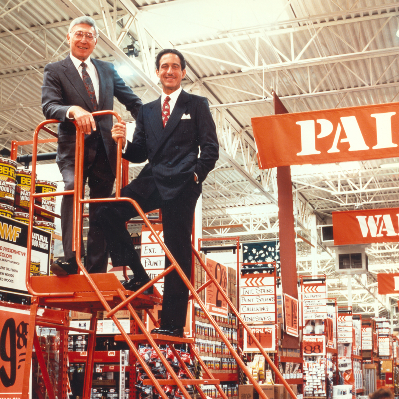 Bernie Marcus and Arthur Blank posing on a ladder inside a Home Depot store