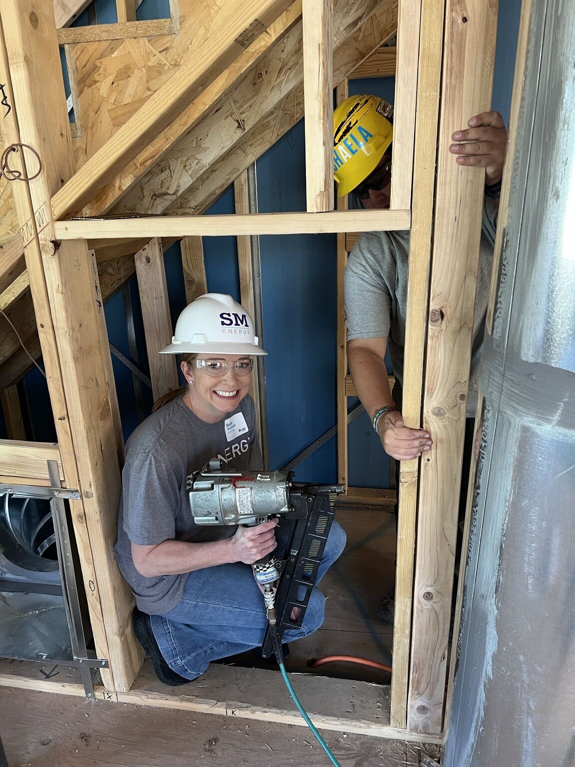 Beth McDonald in safety glasses and a hard hat, holding a nail gun inside a house under construction