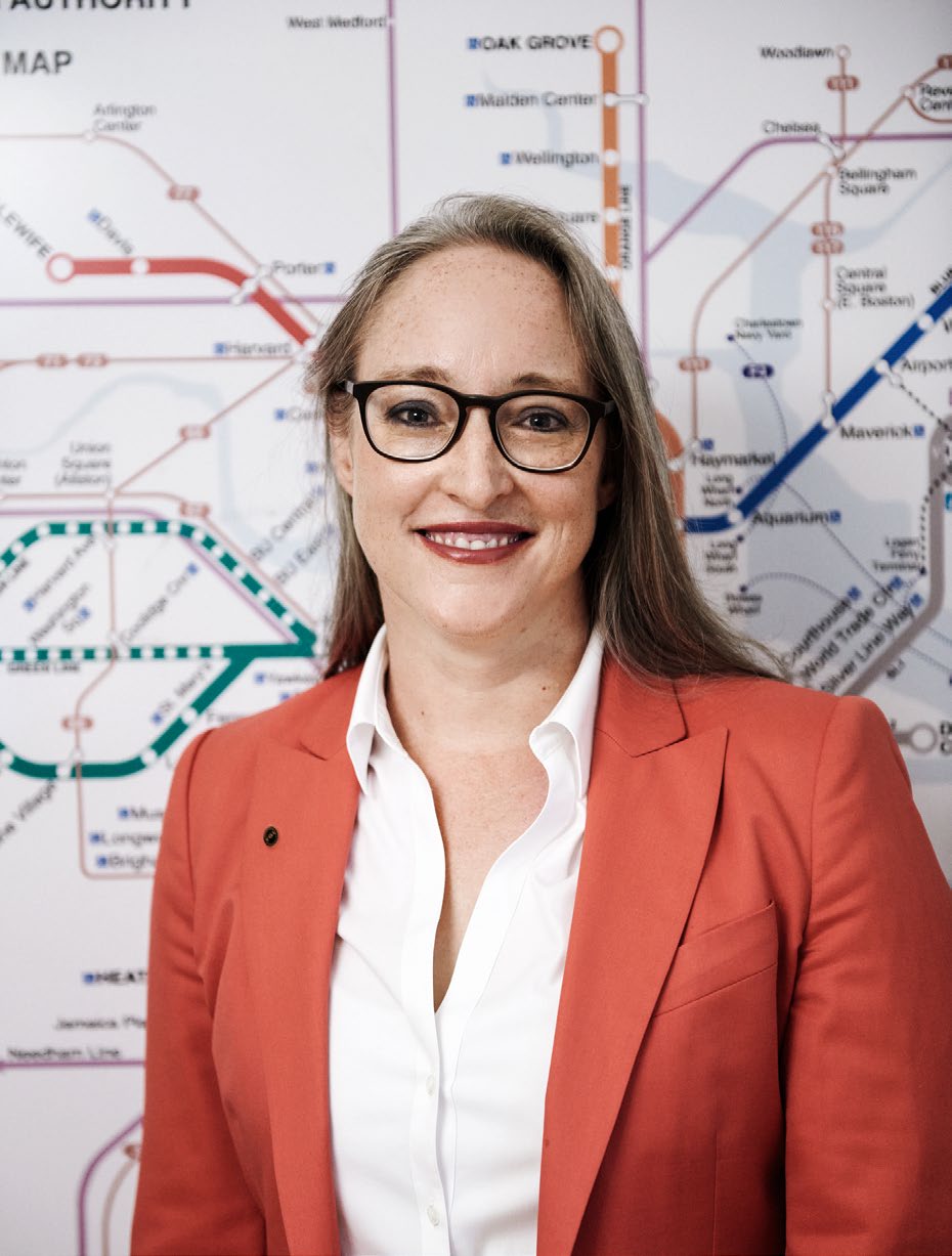 Bettina Hein in a coral jacket, standing in front of a map of Boston’s transit system