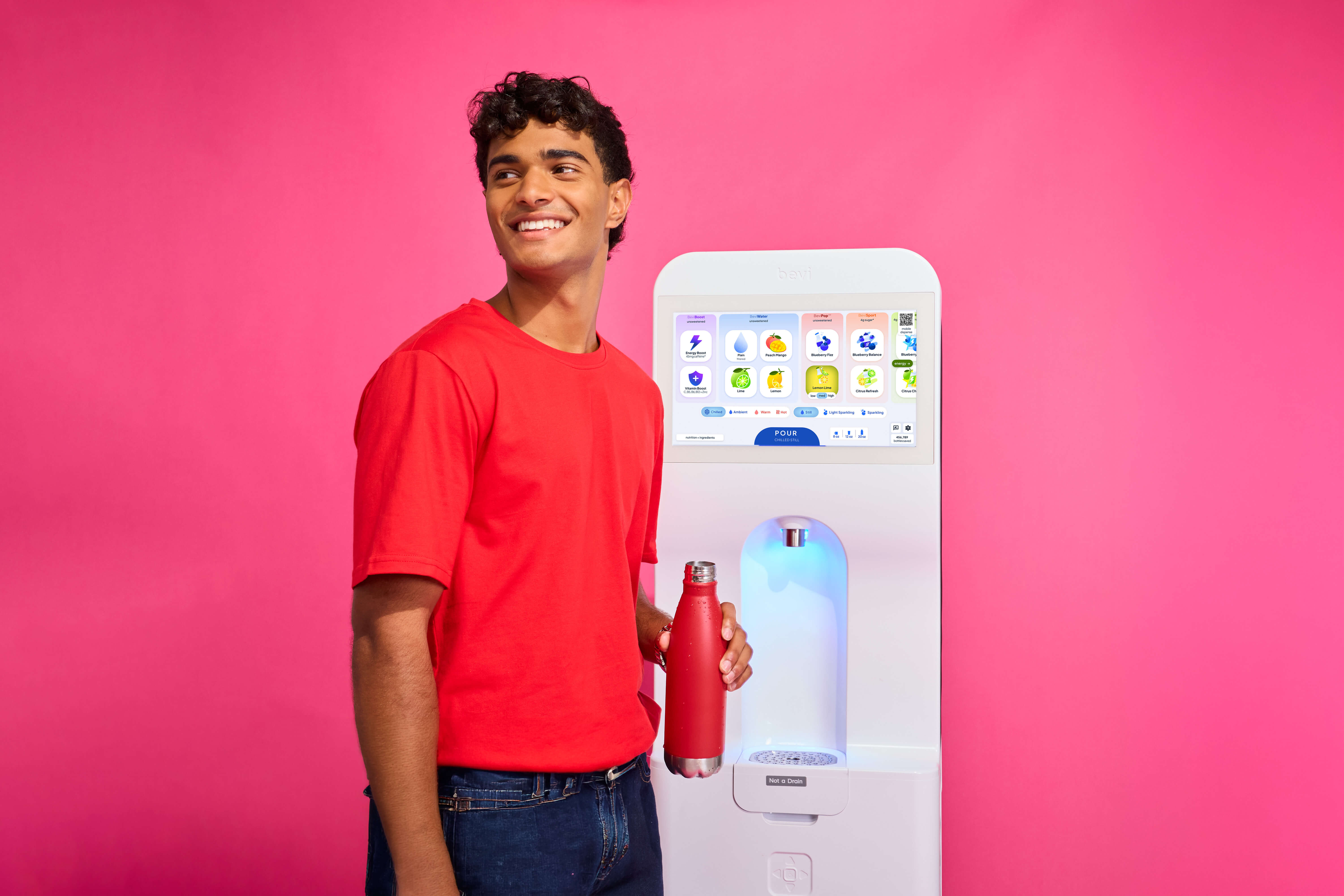 A person holding a reusable bottle beside a Bevi freestanding dispenser against a pink studio backdrop