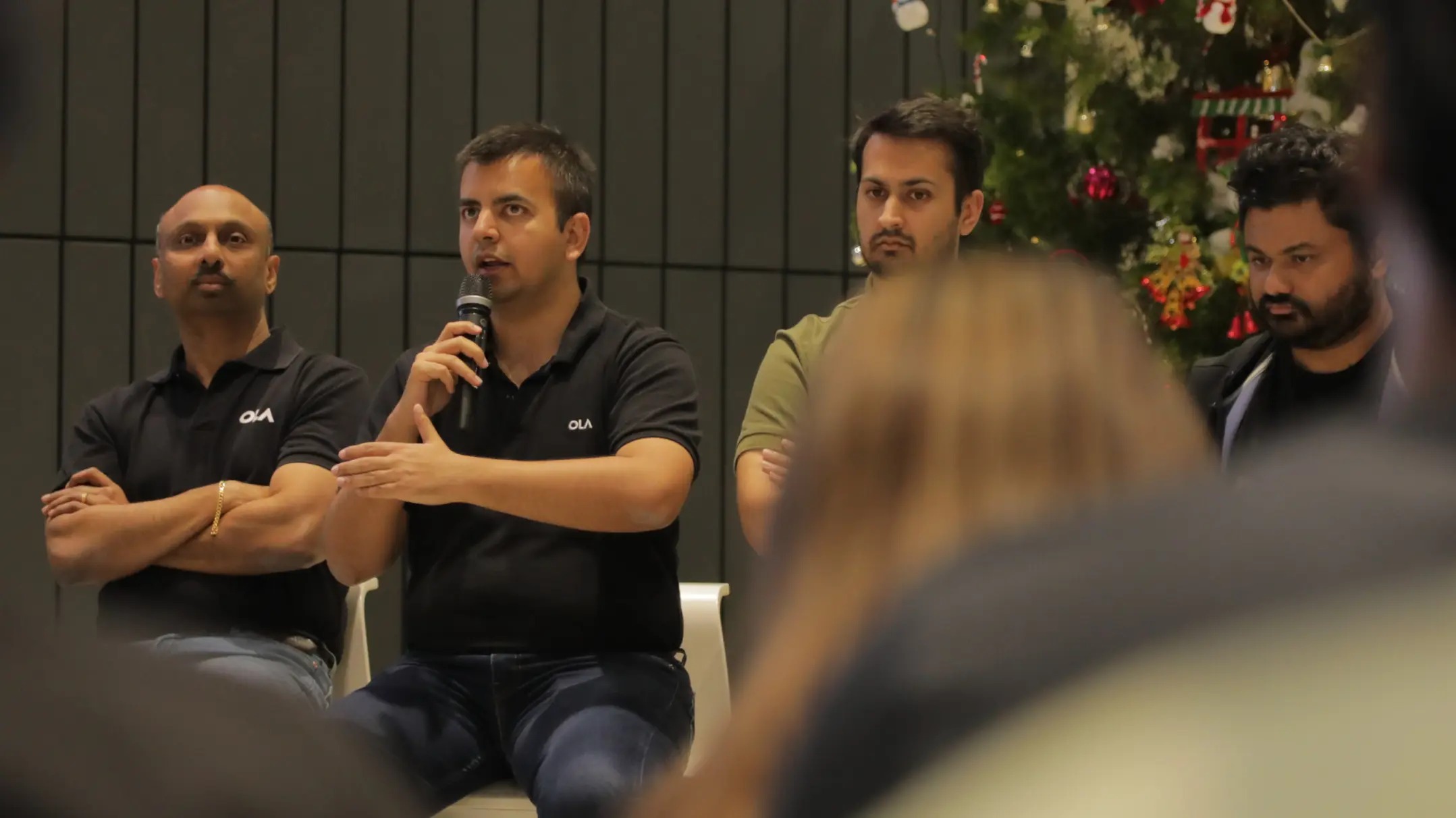 Bhavish Aggarwal speaking into a microphone at an Ola event with colleagues beside him