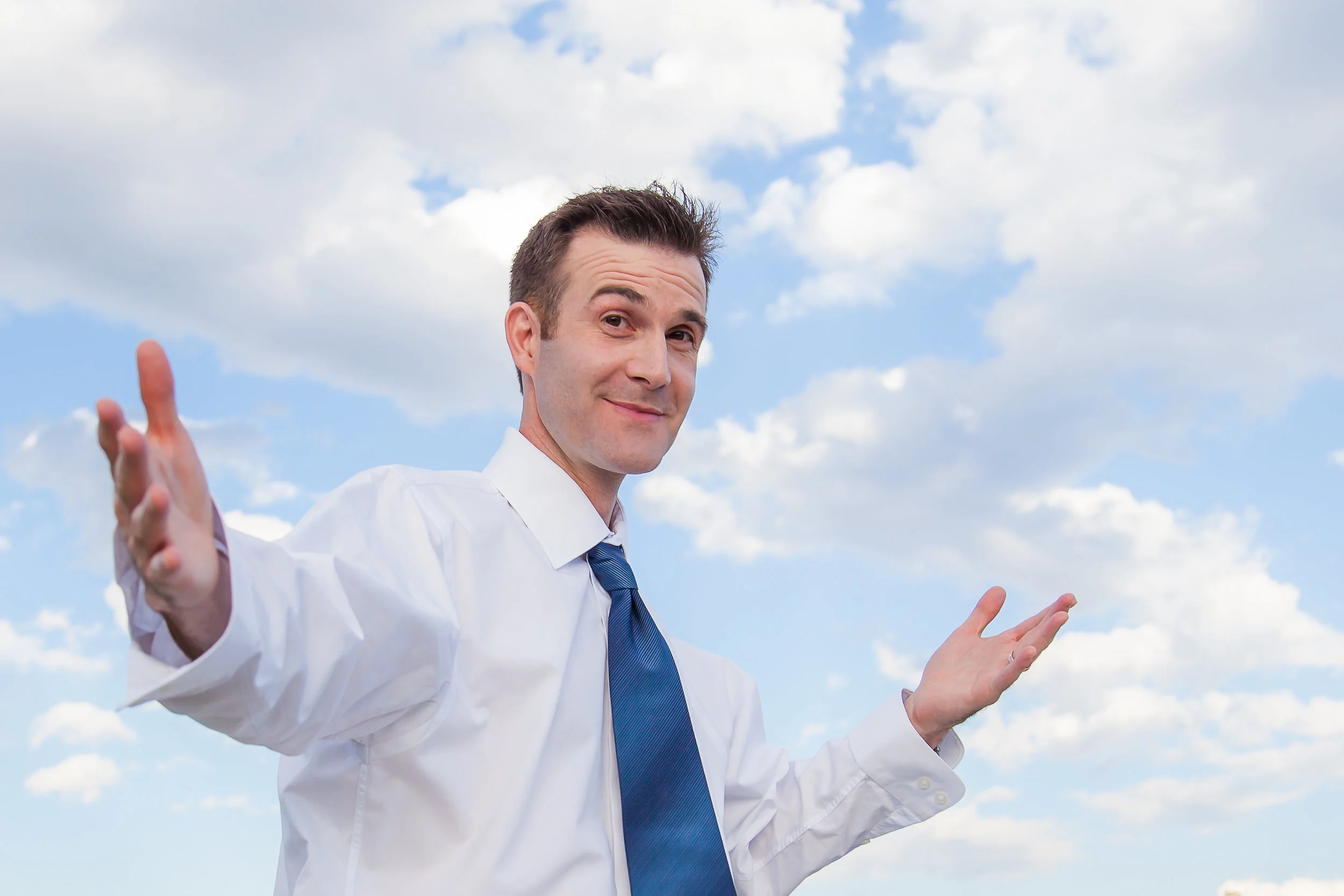 BiggerPockets founder Joshua Dorkin outdoors, wearing a white shirt and blue tie, with his arms extended.