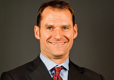 Bill Szlaius in a dark suit, blue shirt and patterned red tie in his 2015 press portrait
