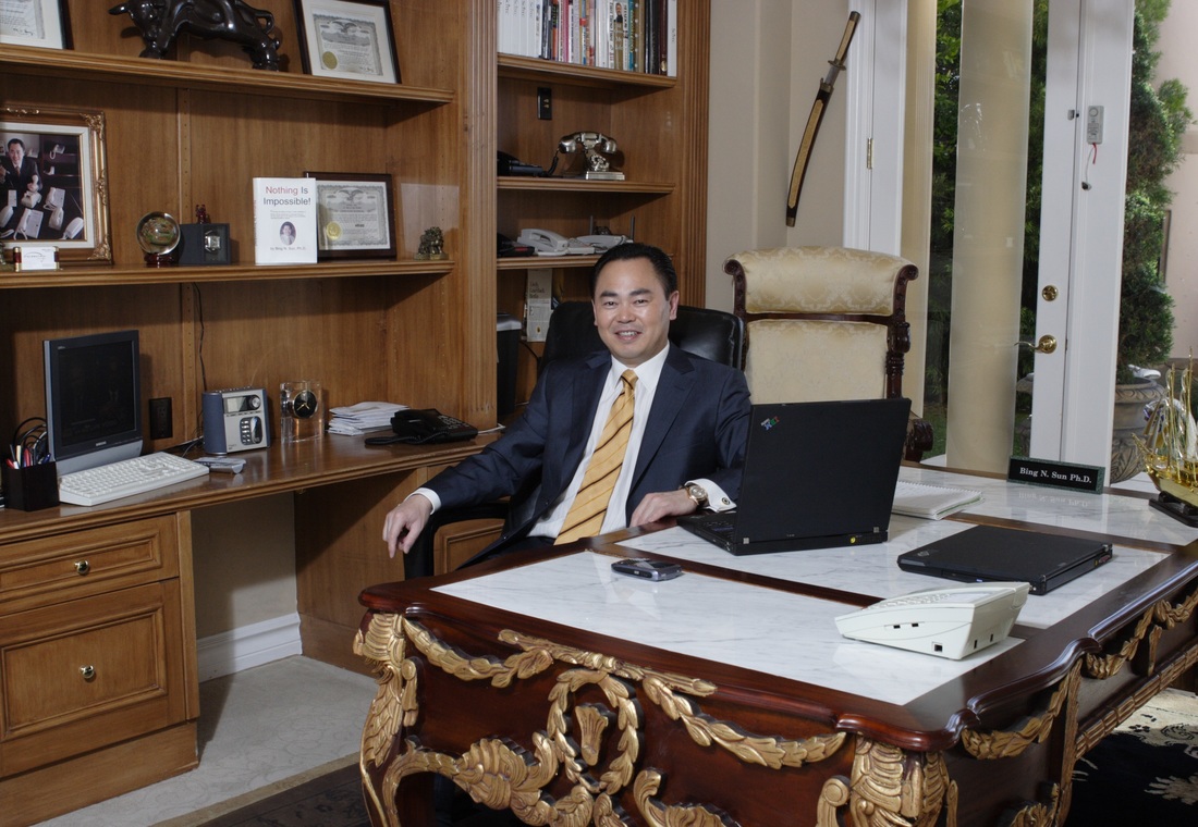 Bing N. Sun seated at his desk in a wood-paneled home office, with telephones nearby